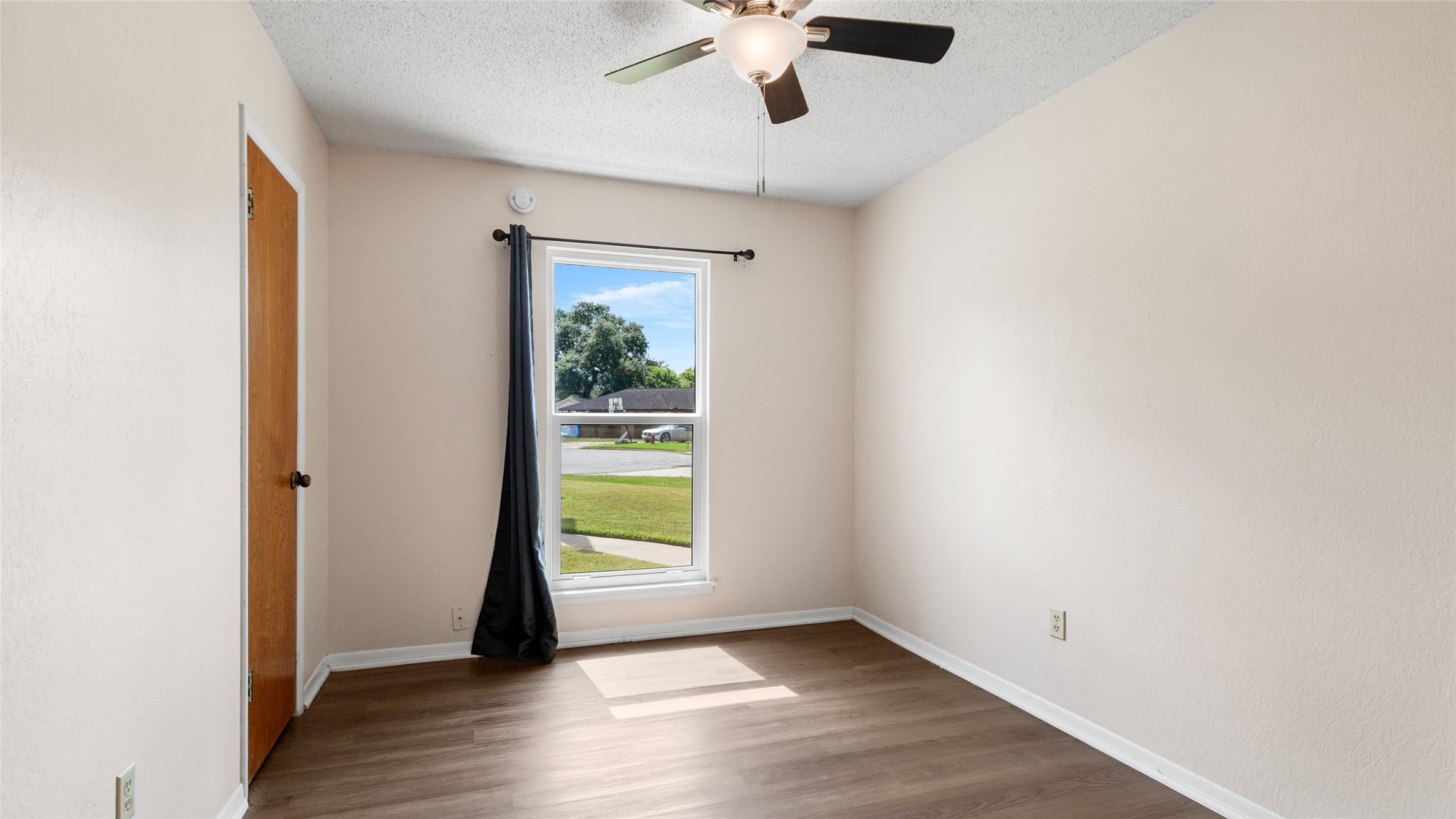 Bright secondary bedroom with wood-look flooring, ceiling fan, closet, and a large window providing abundant natural light.