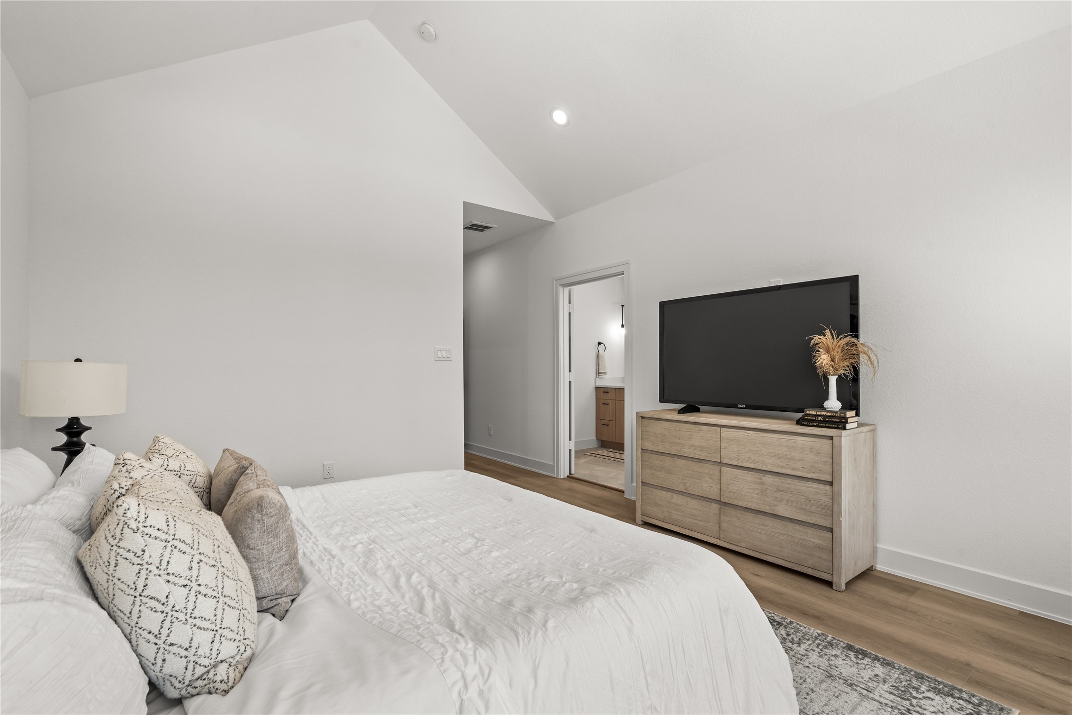 This view from the windows shows the smooth transition from the bed to the rest of the suite. The wide layout provides a clear path toward the attached primary bathroom, while a low-profile media console opposite the bed keeps the room clean and clutter-free.