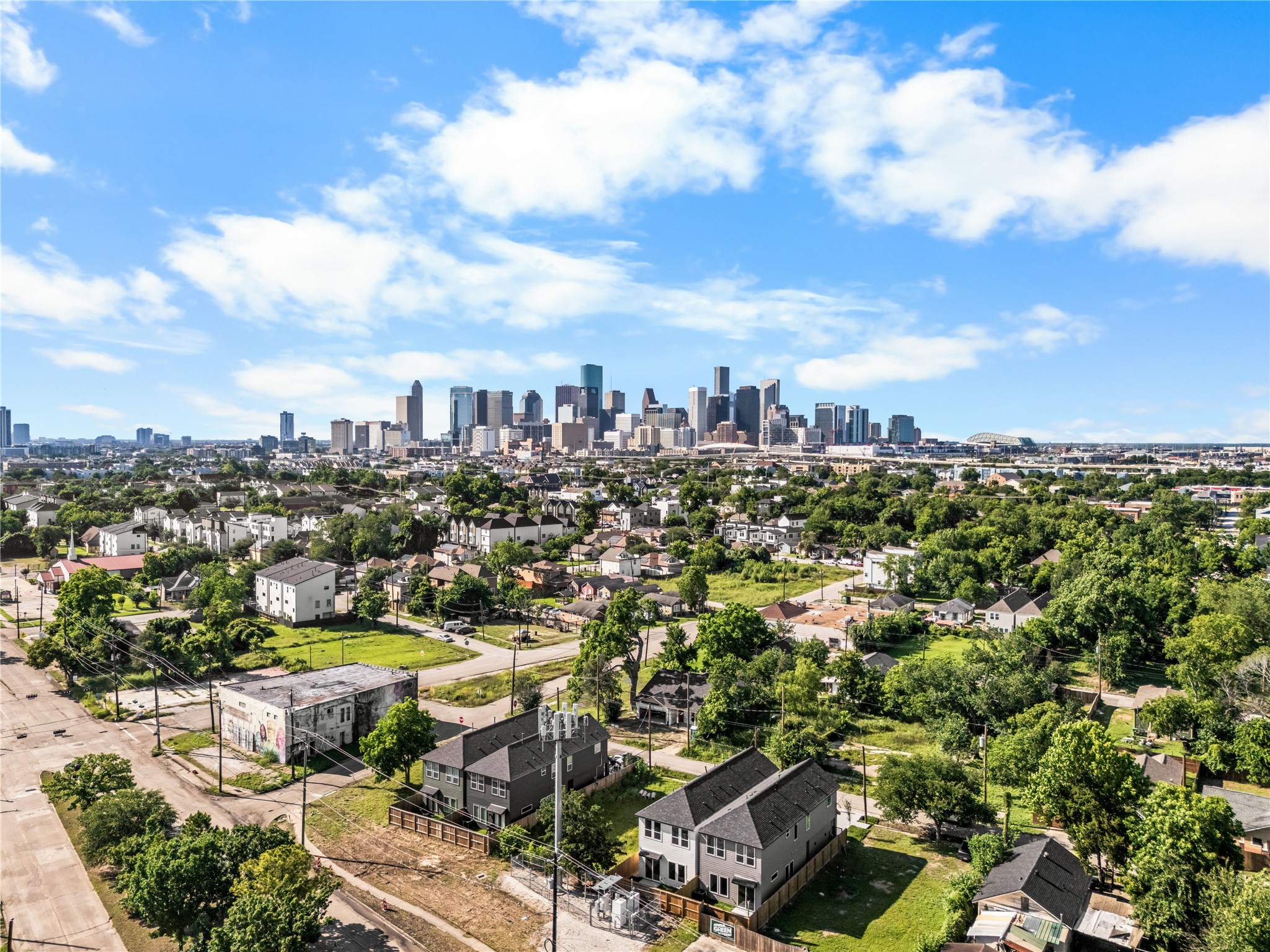 This drone view shows the home's proximity to downtown, nestled in a residential neighborhood surrounded by mature trees. Under a clear blue sky, the property offers an unobstructed view of the city skyline, highlighting its balance of neighborhood charm and urban access.