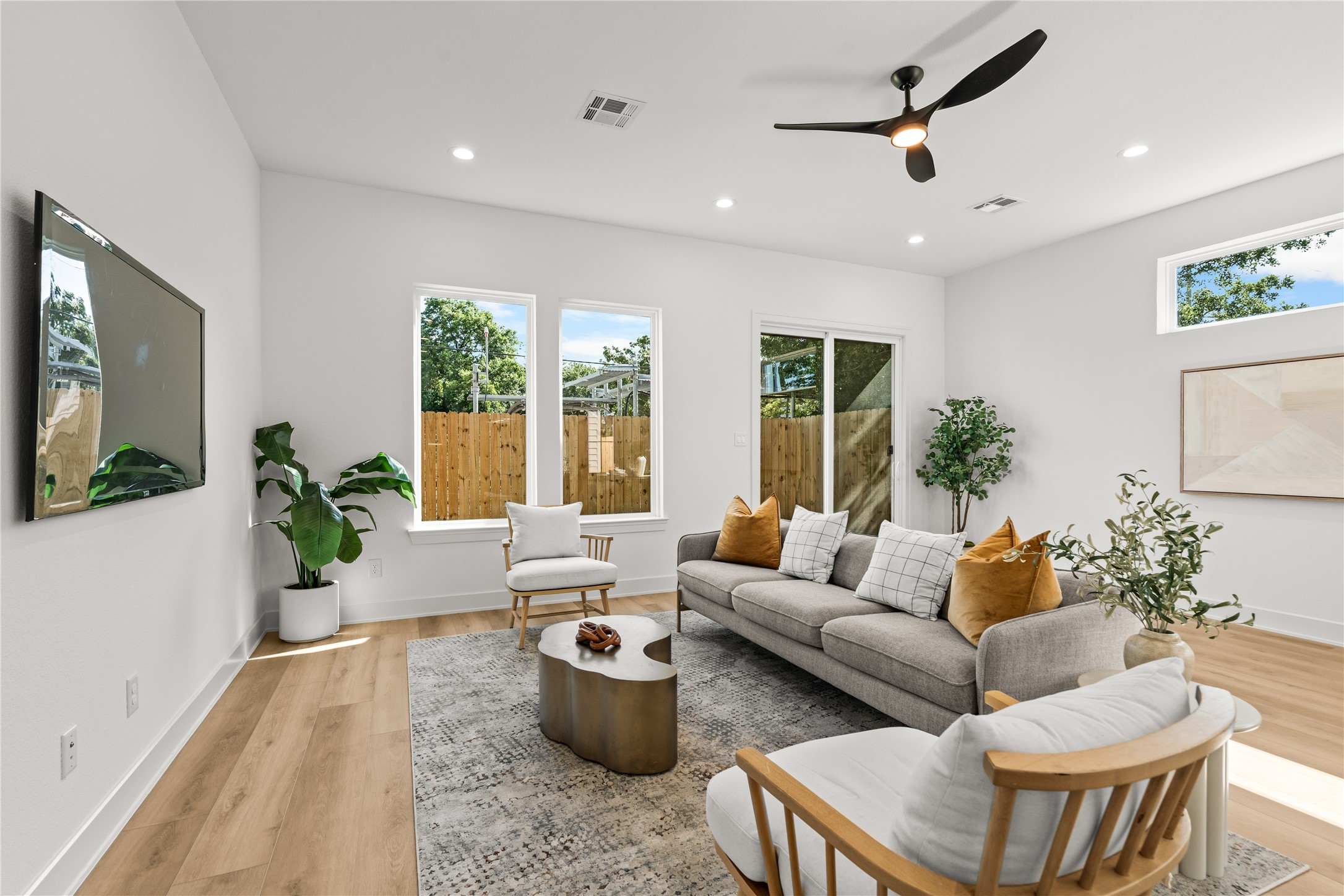 The main living room is bright and open, featuring a modern/spacious layout. Large windows look out into the backyard, providing a clean view of the horizontal wood fence and greenery while letting in plenty of natural light.