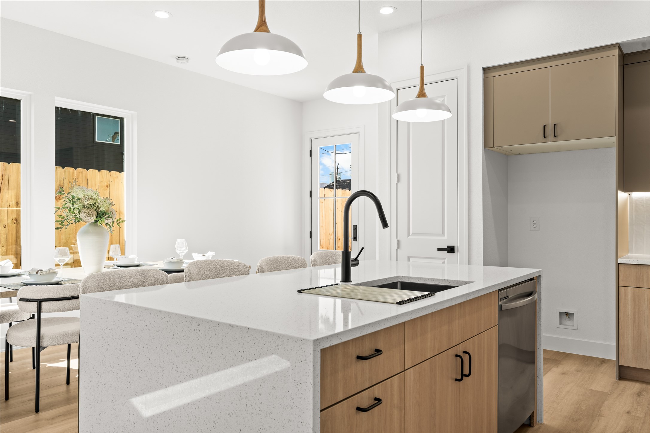 The kitchen island features a white quartz countertop with a deep, under-mount stainless steel sink and a matte black pull-down faucet. A stainless steel dishwasher is built right into the natural wood lower cabinets directly across from the sink, making cleanup easy.
