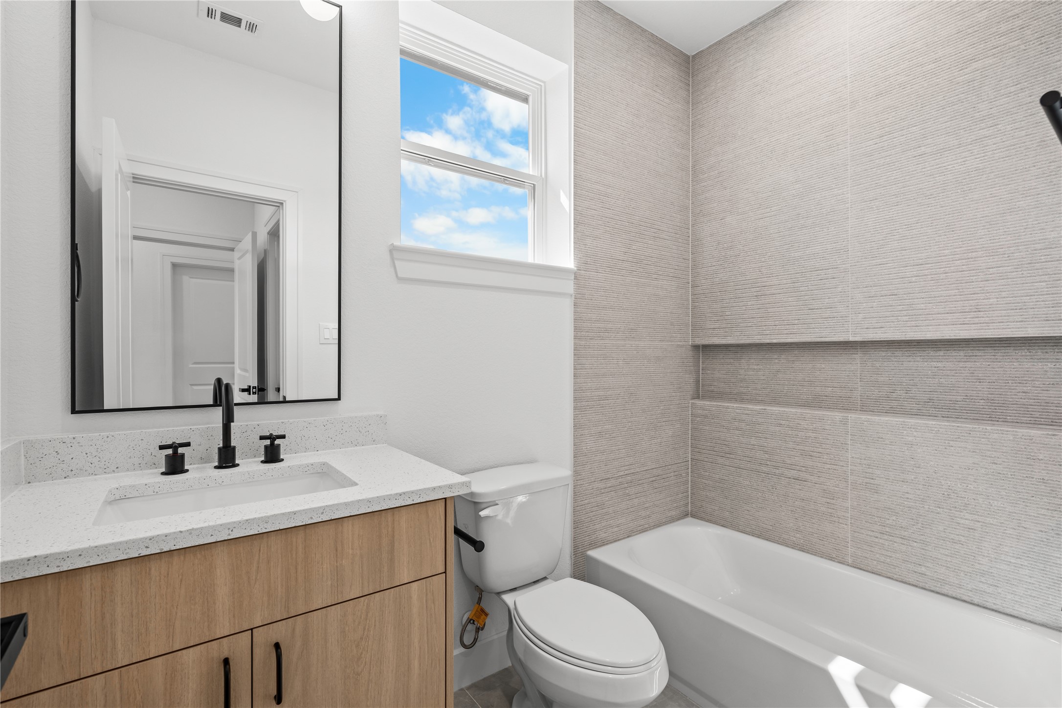 The third full bathroom features a warm wood vanity, a white quartz countertop, and a shower-tub combination. A large vertical window brings in excellent natural light, complemented by a durable, textured tile surround.