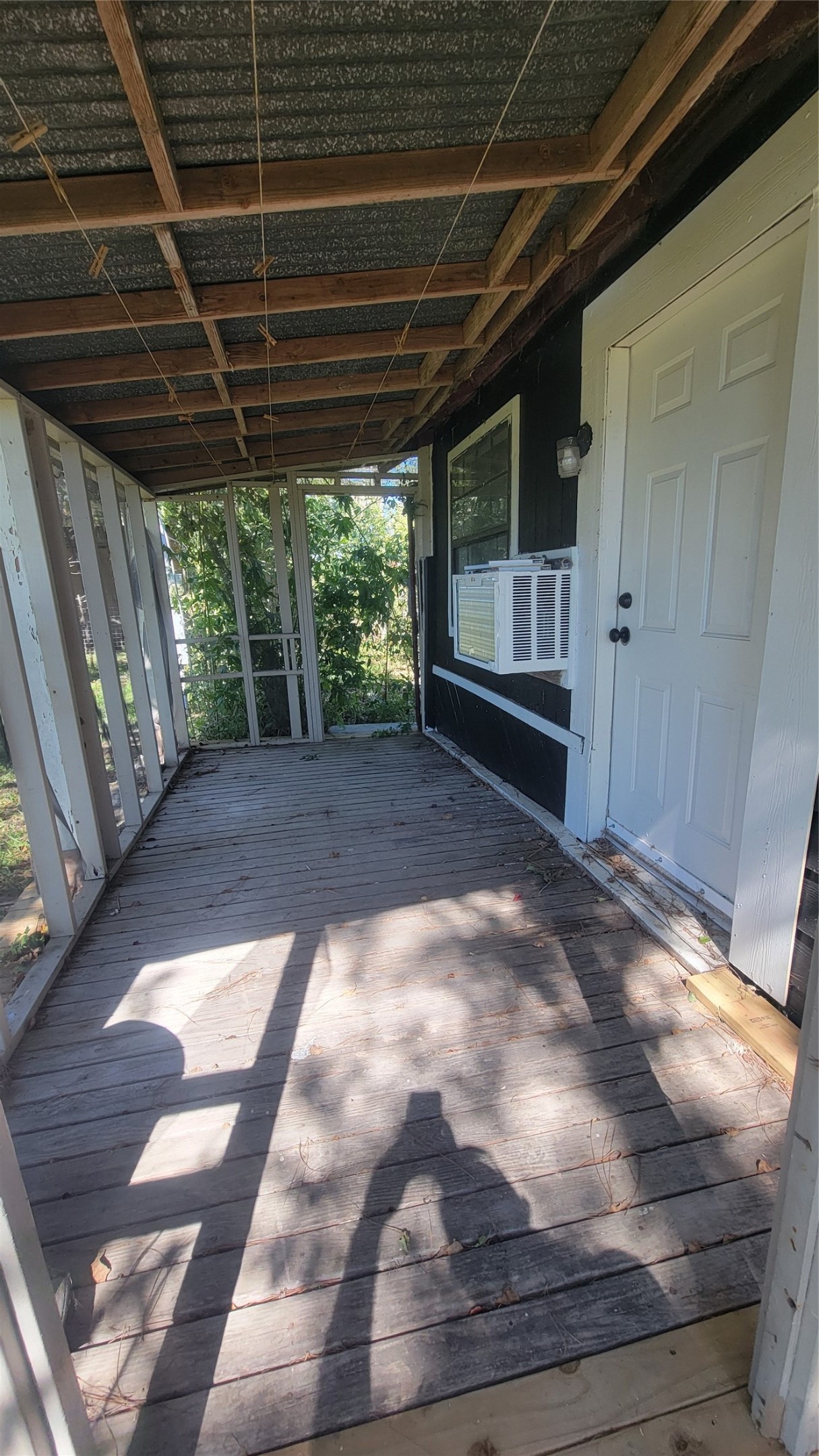 This photo showcases a rustic, covered porch with a wooden floor and open railing, featuring a side door and a window AC unit. The porch is shaded, providing a cozy outdoor space.