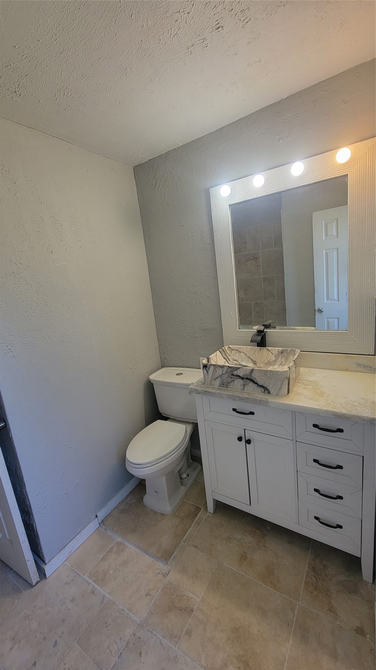 This bathroom features a modern design with a white vanity and marble vessel sink. It has a large, lighted mirror and neutral tile flooring, creating a clean and bright space.