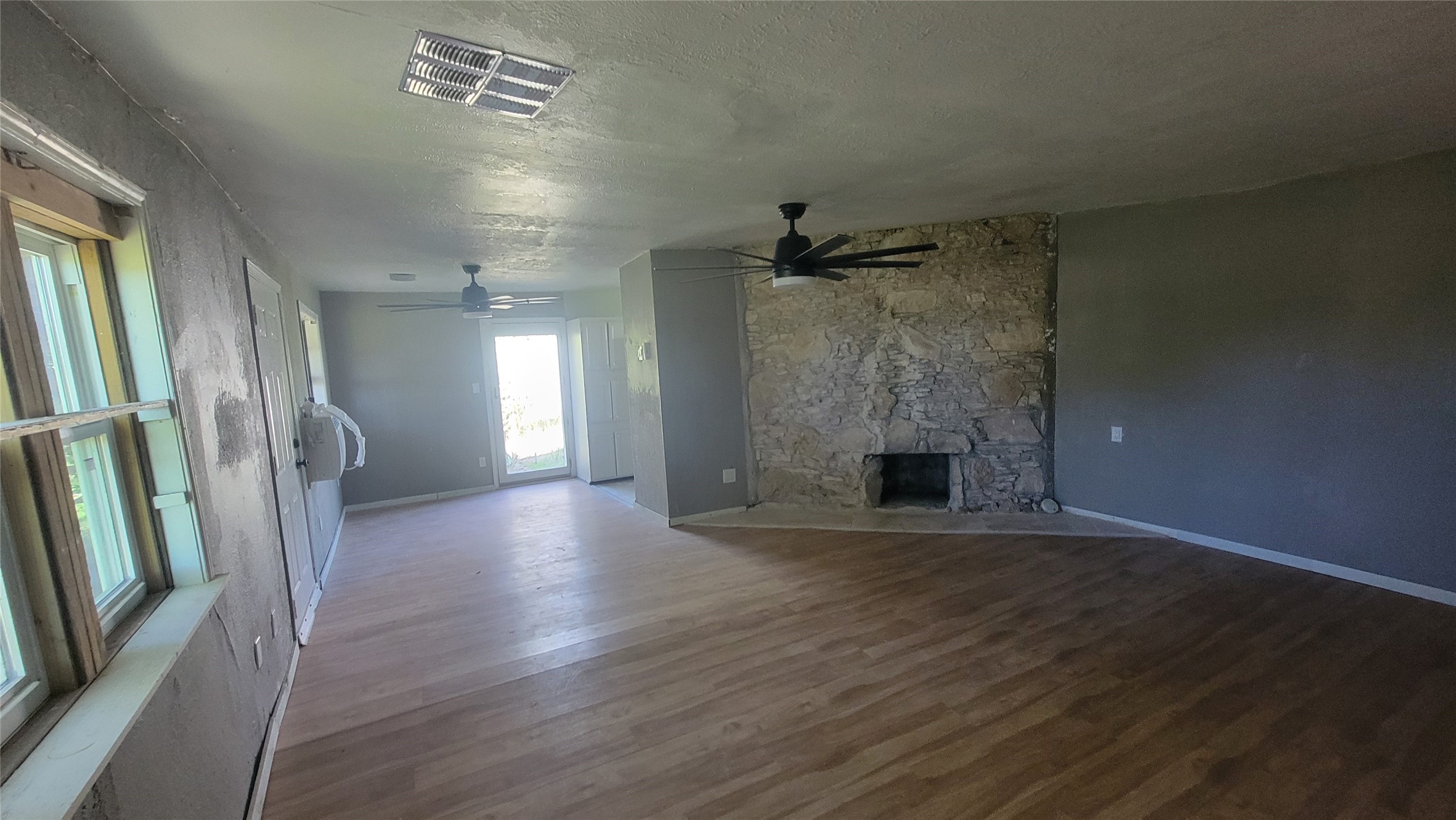 This spacious living area features wood flooring, a stone fireplace, and ceiling fans. Large windows and a glass door provide plenty of natural light, creating an inviting atmosphere.