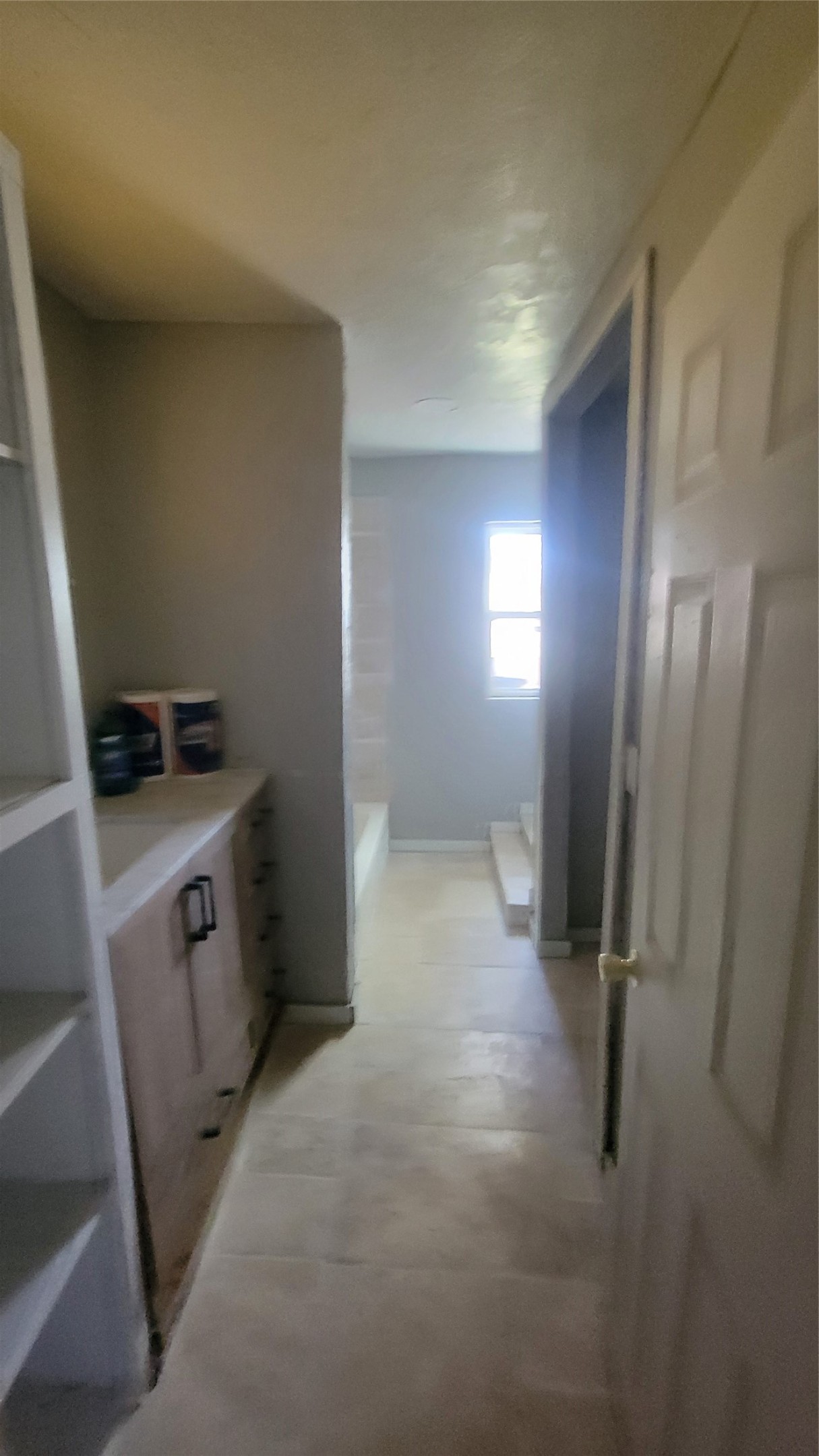 This photo shows a narrow bathroom with beige tile flooring and a window providing natural light. The room includes built-in shelving on the left and a wooden vanity with storage. The bathtub is partially visible at the end of the room.