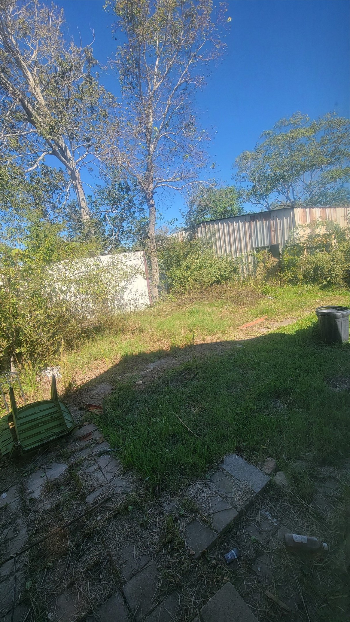 This photo shows a backyard with a grassy area, some overgrown vegetation, and a few trees. There are pavers on the ground and a fallen outdoor chair. A metal fence is visible in the background, providing some privacy.
