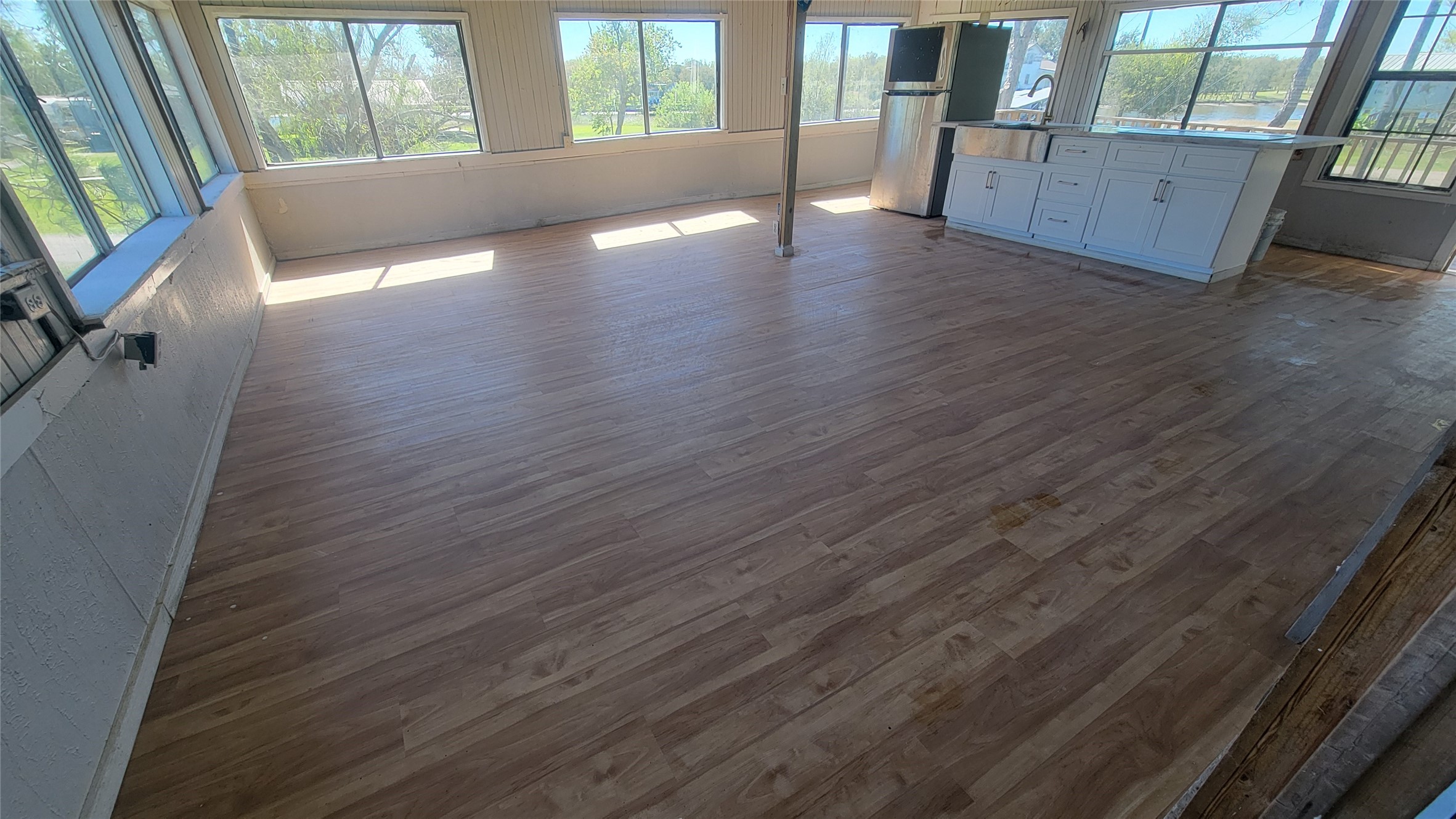This bright, open-concept room features expansive windows for natural light, hardwood flooring, and a modern kitchen area with white cabinetry and a small island. Ideal for entertaining or family living.