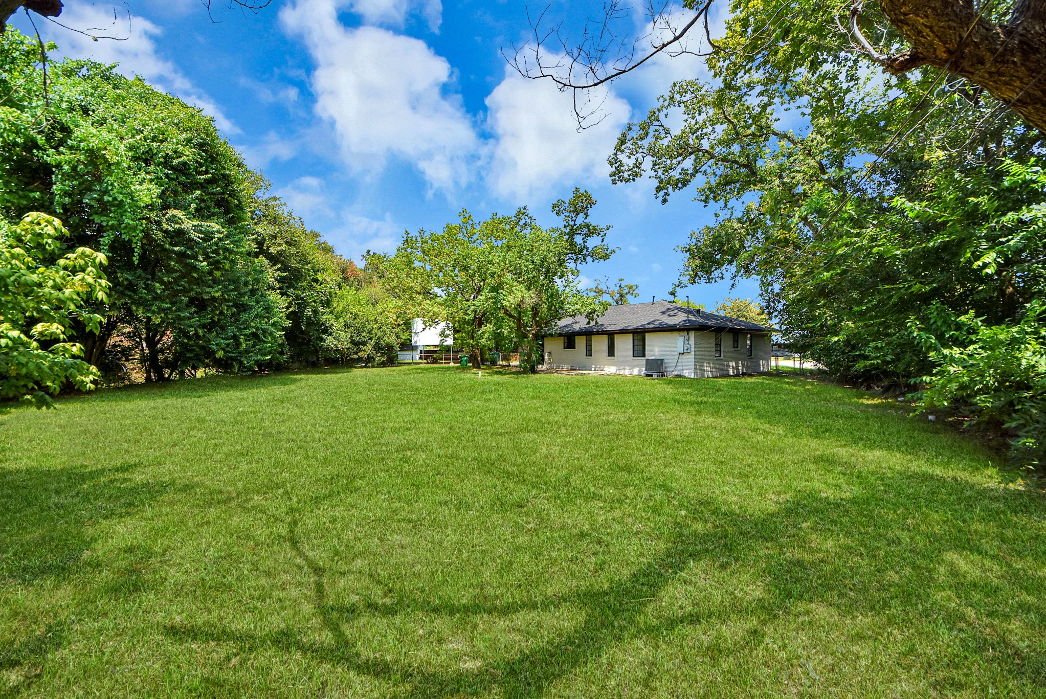 Expansive green backyard surrounded by mature trees, offering privacy and tranquility with a cozy single-story home nestled in the corner.