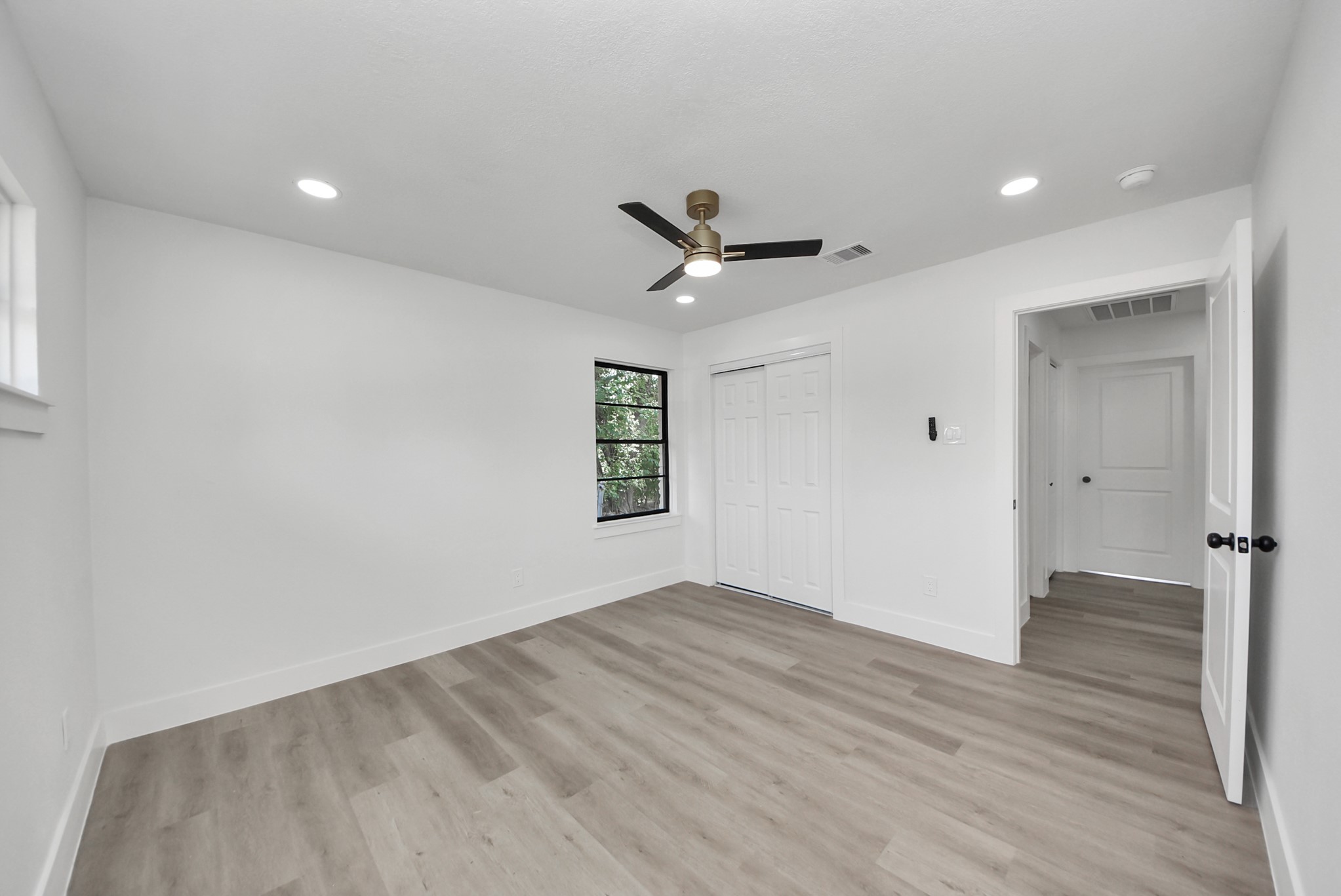 Spacious and bright room featuring modern recessed lighting, sleek ceiling fan, and elegant wood flooring, with ample closet space and a large window for natural light.