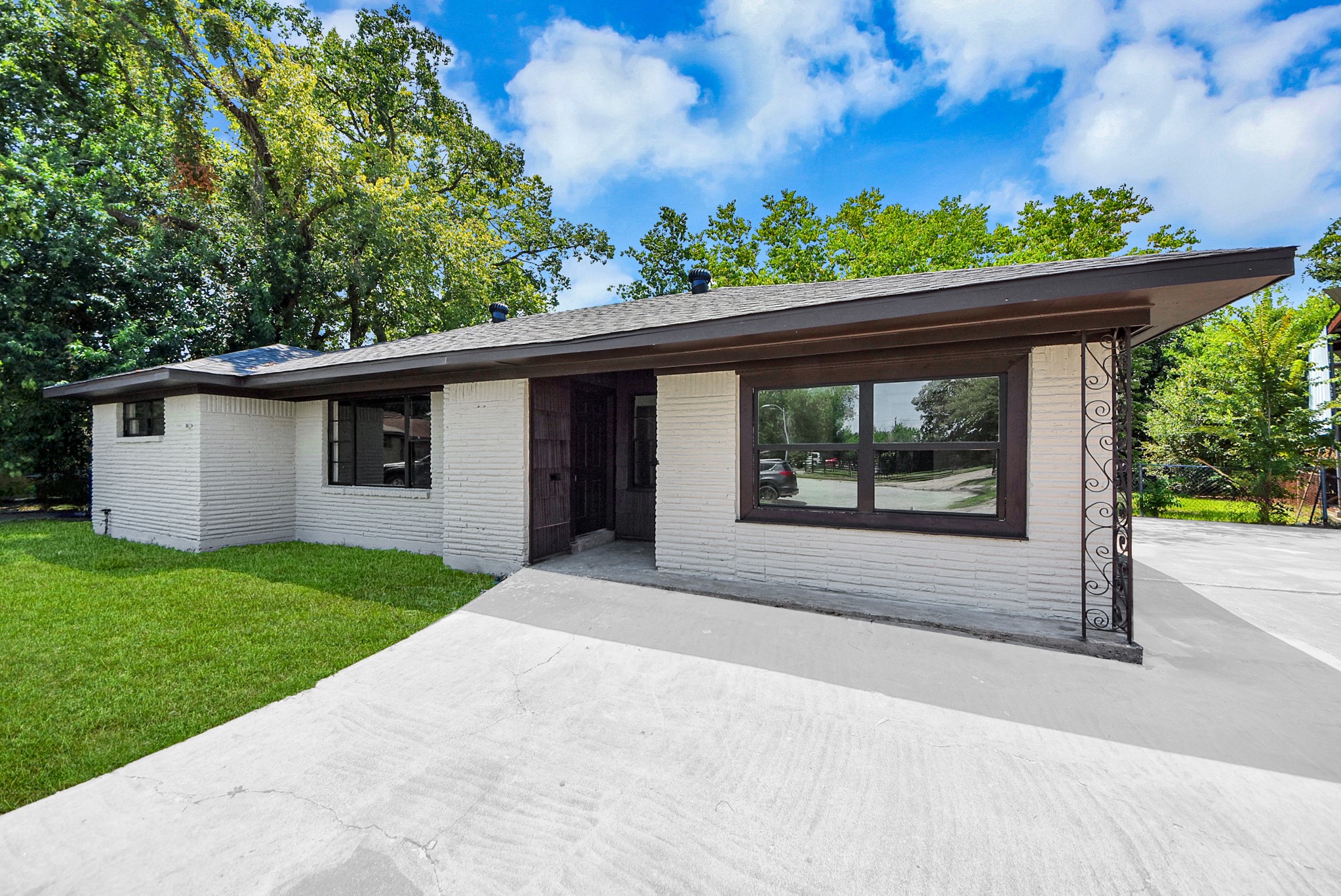 Charming single-story home with a sleek white brick exterior, large windows, and a spacious driveway, set against a backdrop of lush greenery.