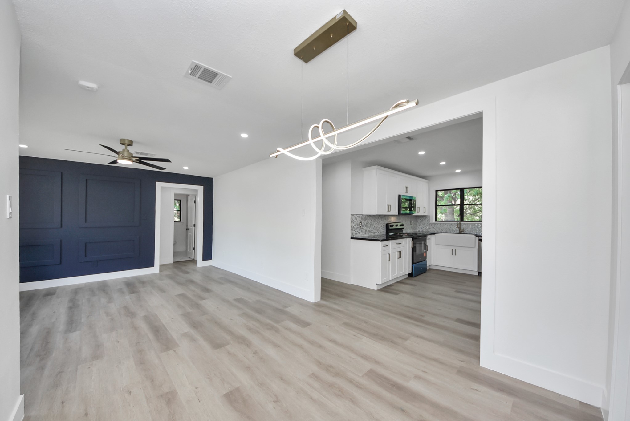 Experience modern elegance in this open-concept living space featuring sleek wood flooring, a striking navy accent wall, and a contemporary light fixture. The adjoining kitchen is beautifully appointed with white cabinetry and stylish backsplash, perfect for entertaining.