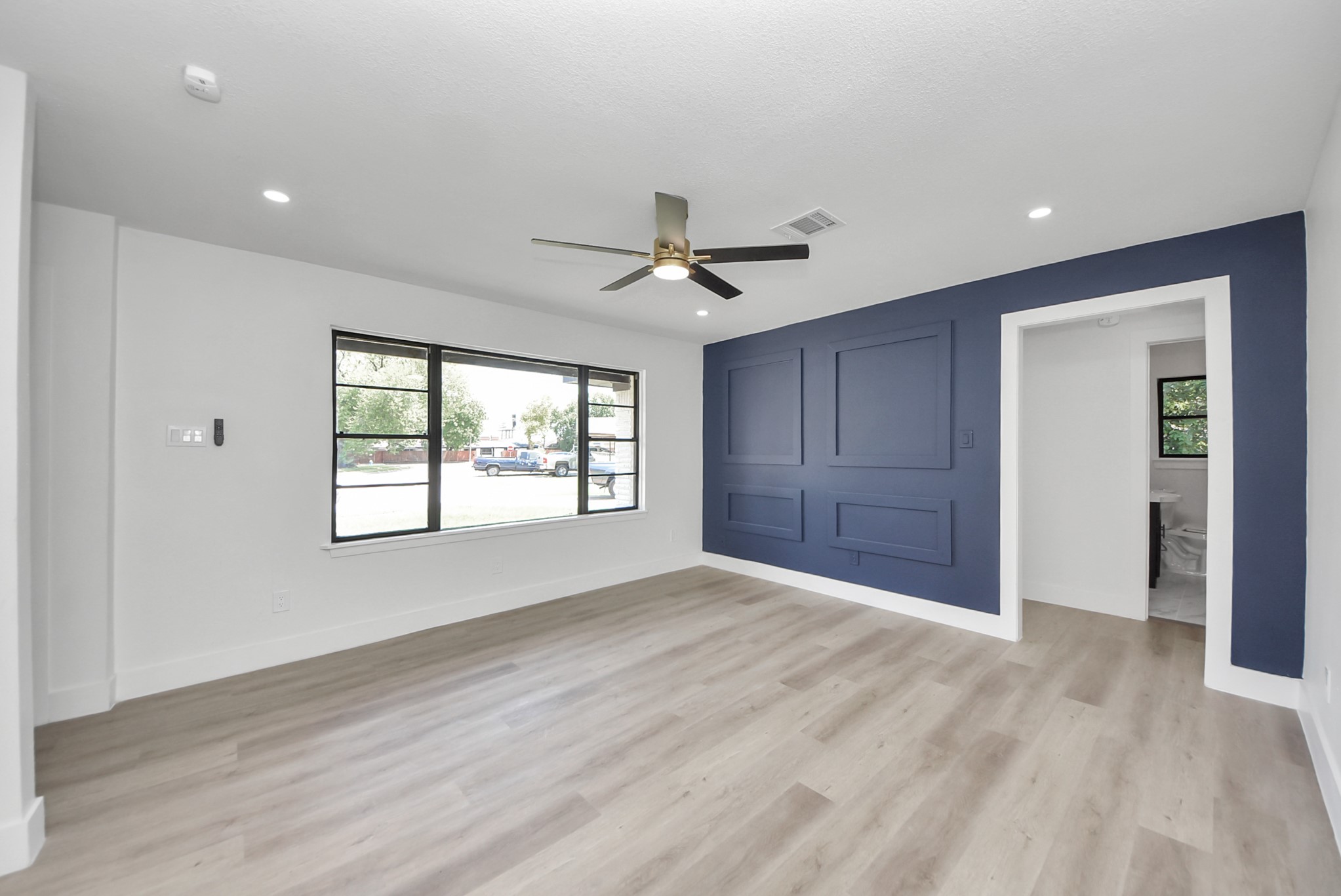 Bright and modern living area featuring a stylish accent wall, large windows, and sleek wood flooring, complemented by a contemporary ceiling fan.