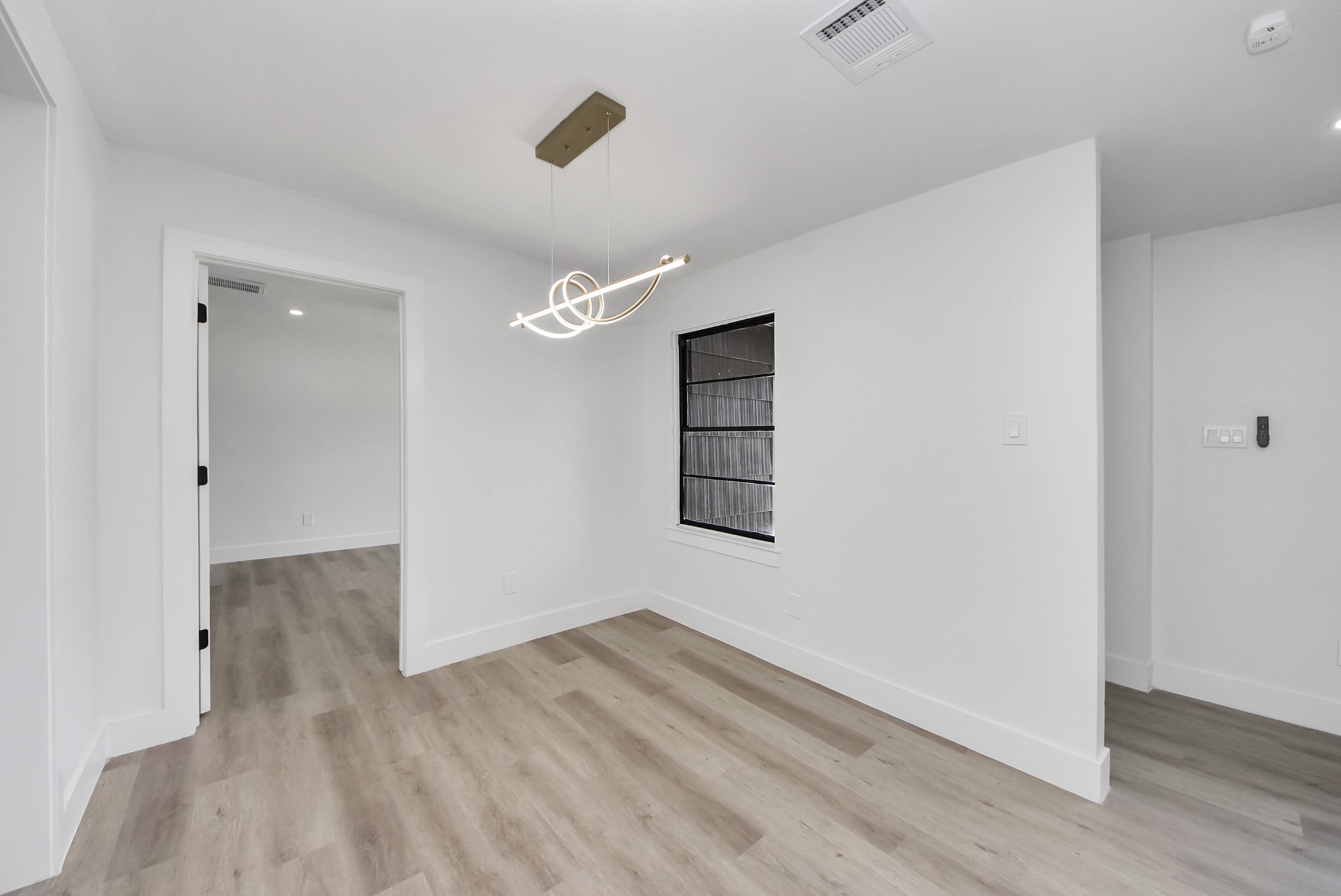 Bright and modern room featuring sleek wood flooring, a contemporary light fixture, and a large window, offering a versatile space for dining or lounging.