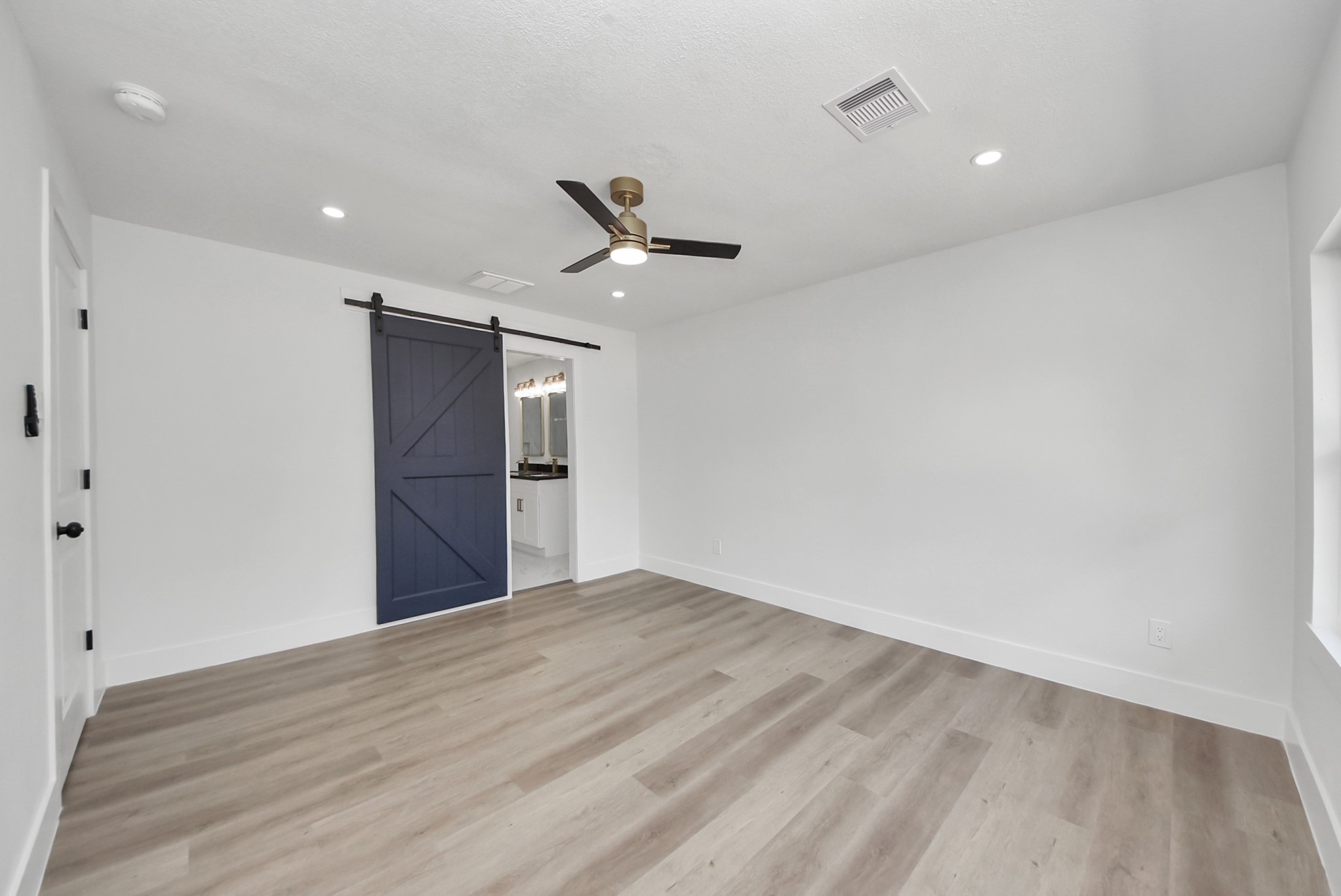 Spacious room with elegant wood flooring, modern recessed lighting, and a stylish ceiling fan, featuring a striking blue barn door leading to a well-lit bathroom.