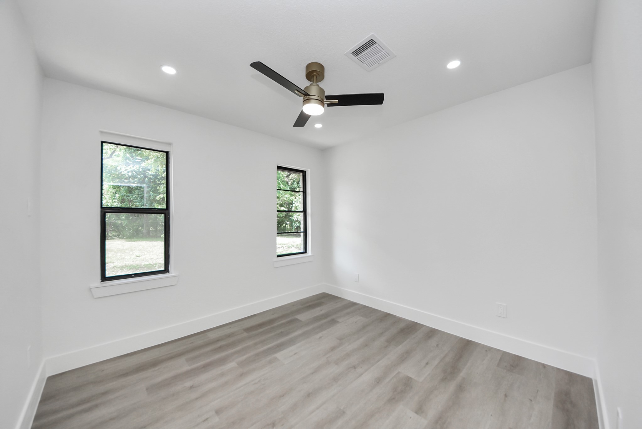 Bright and airy room featuring sleek laminate flooring, fresh white walls, and large windows offering serene outdoor views, complemented by a modern ceiling fan for comfort.
