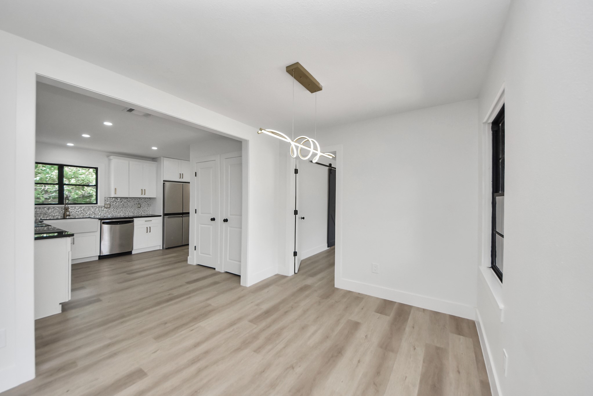 Bright and modern space with sleek light wood flooring, a contemporary light fixture, and an open view into a stylish kitchen featuring stainless steel appliances and a farmhouse sink.