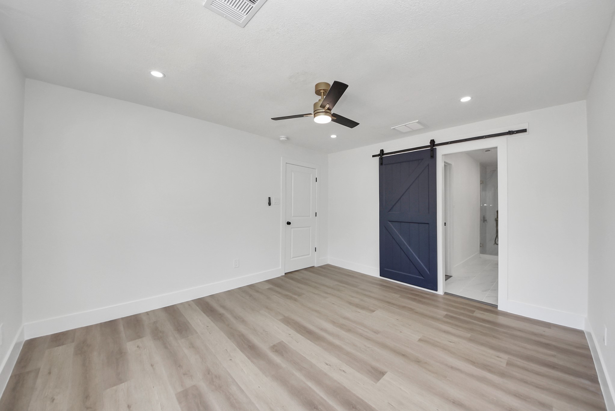 Spacious room with sleek wood flooring, recessed lighting, and a stylish ceiling fan. Features a modern barn door leading to an en-suite bathroom.