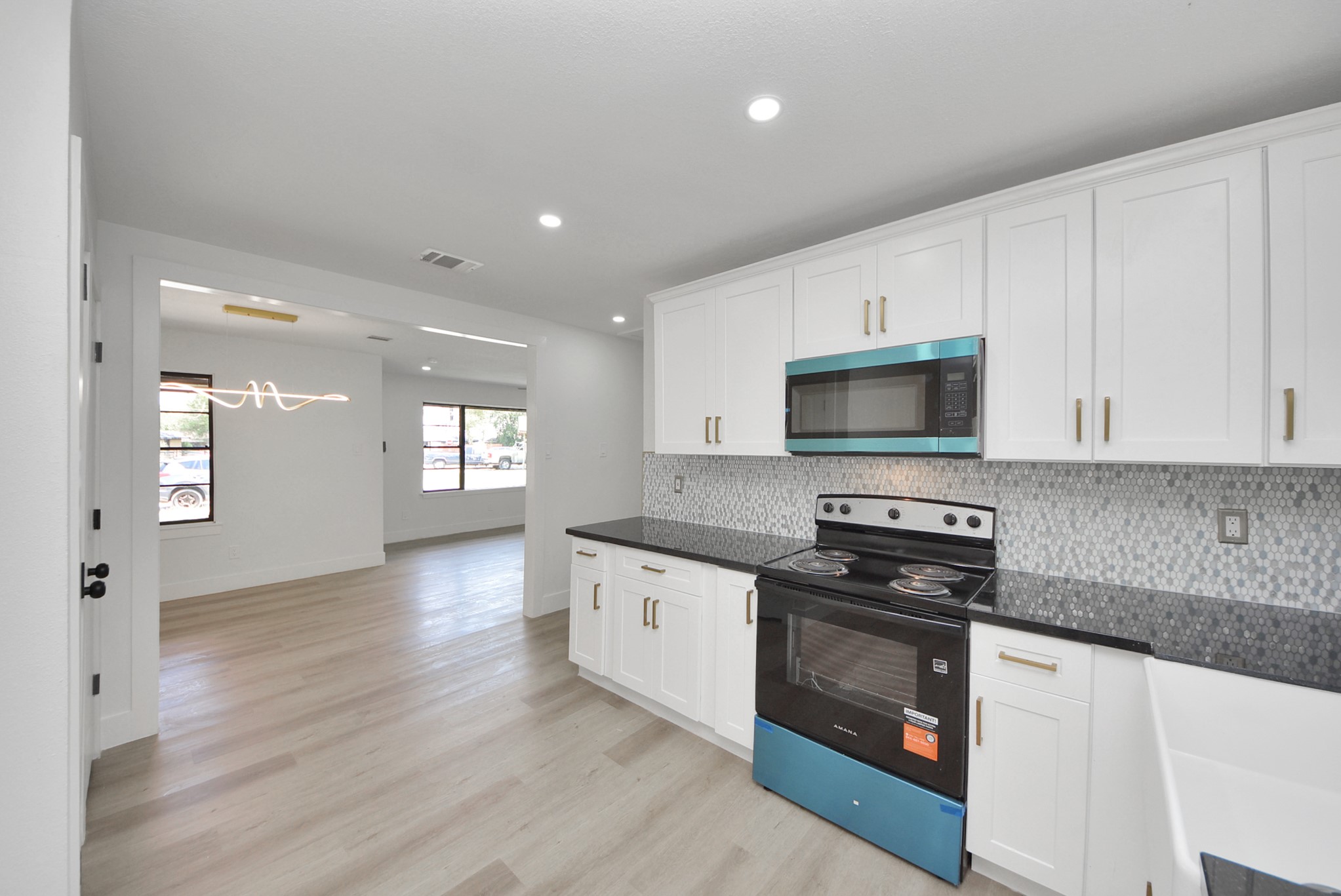 Modern kitchen with sleek black countertops, white cabinetry, and stainless steel appliances, seamlessly flowing into a bright, open living area with large windows.