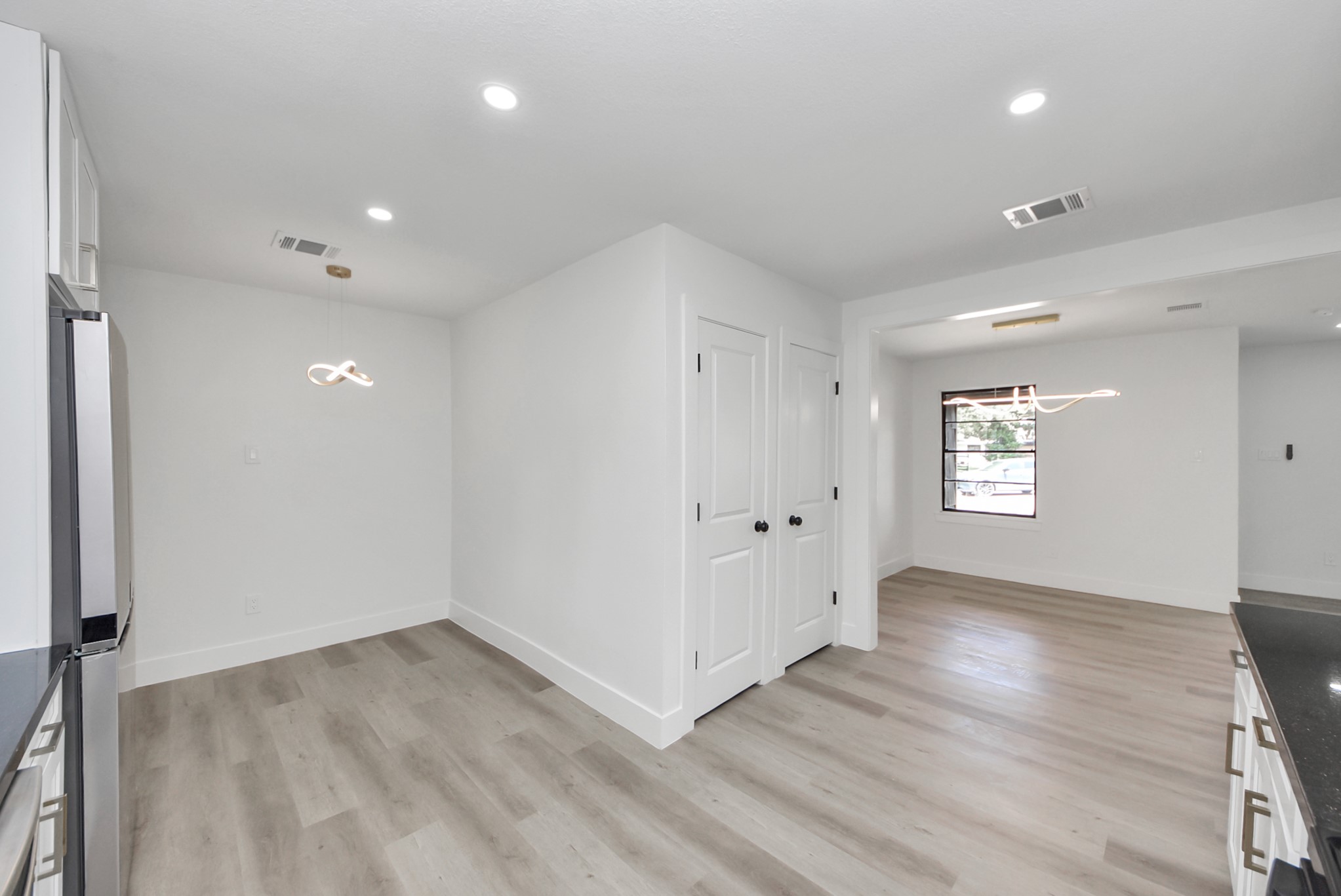 Bright and modern open-concept space featuring sleek hardwood floors, recessed lighting, and a contemporary light fixture. The area is versatile, leading to a window that offers natural light and a view outside.