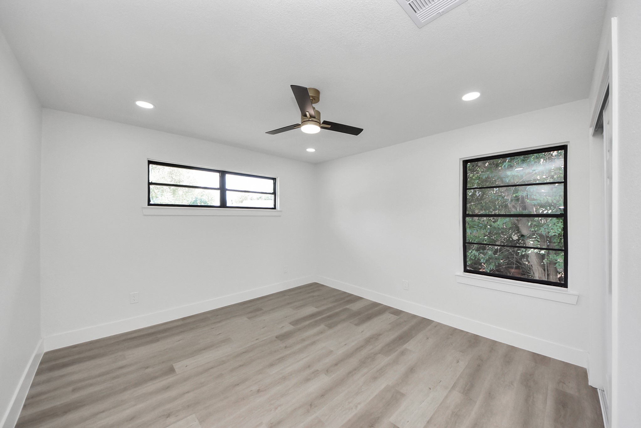Bright, modern room featuring sleek wood flooring, recessed lighting, and large windows with views of lush greenery.