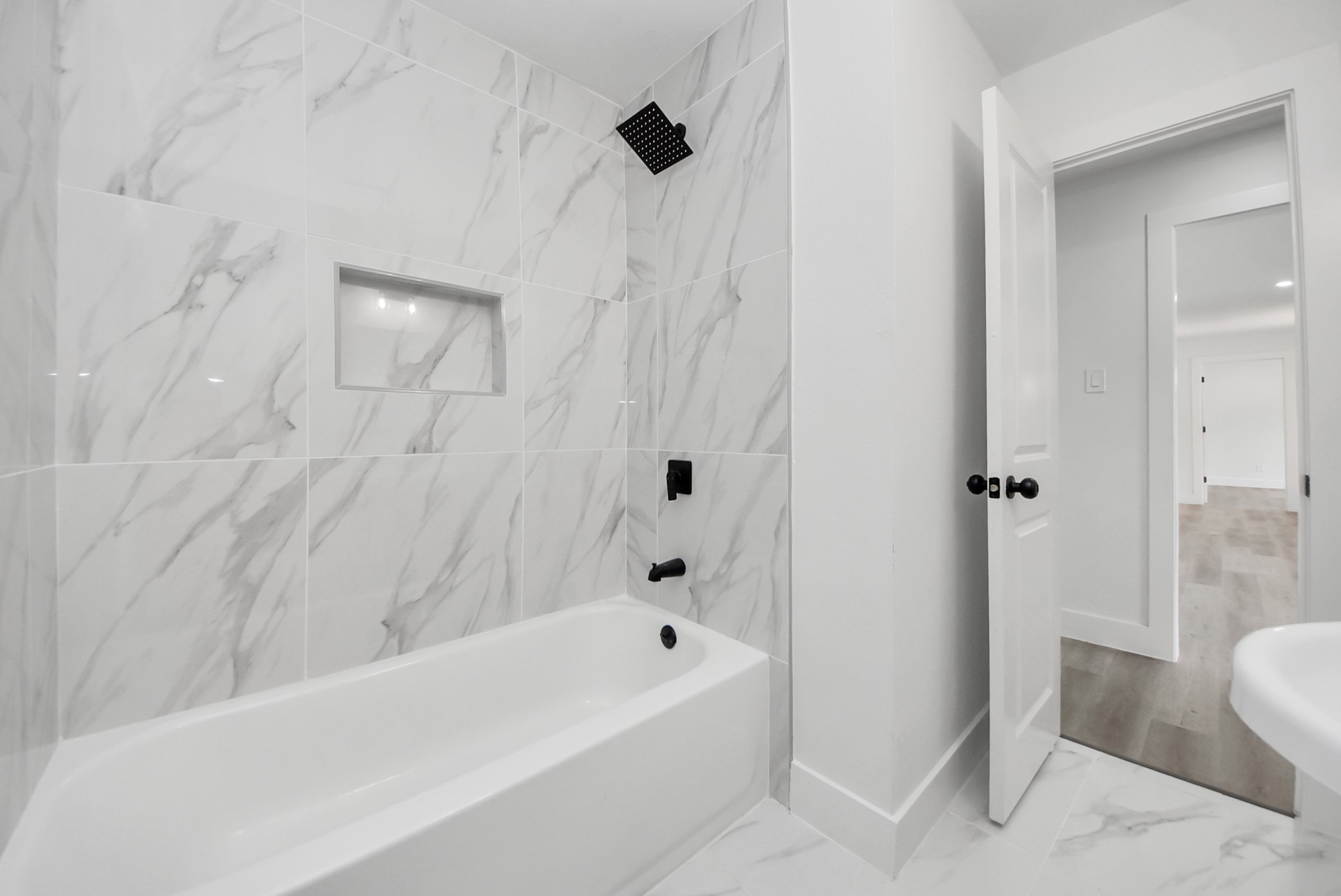 Sleek bathroom featuring elegant marble-patterned tiles, a modern black shower fixture, and a convenient built-in shelf above a pristine bathtub.