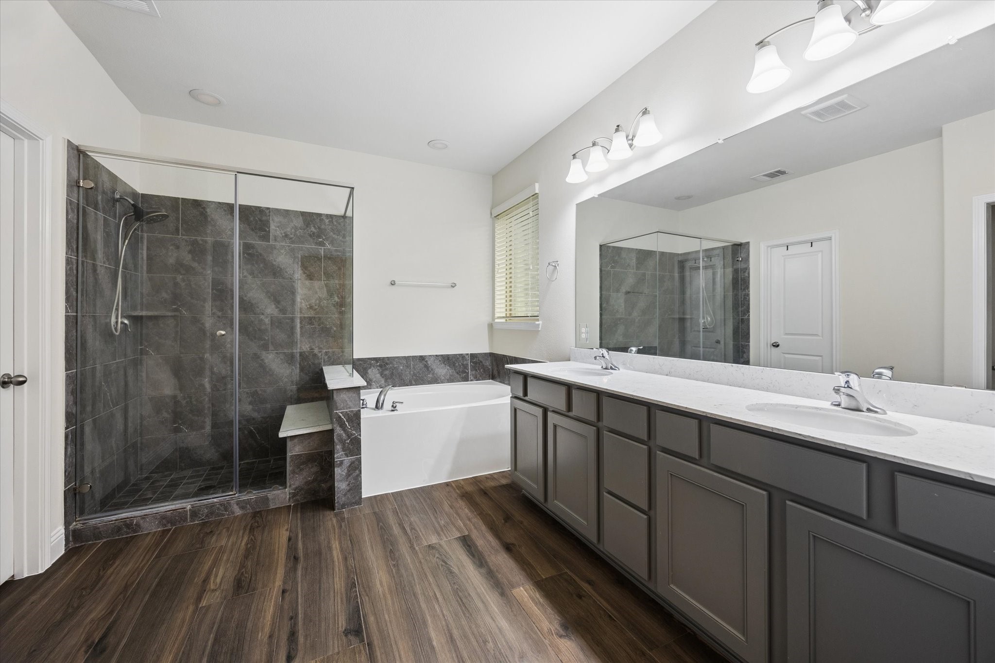 Spacious primary bathroom featuring a large dual-sink vanity with ample storage, a deep soaking tub, and a separate glass-enclosed walk-in shower with elegant tile surround. Neutral finishes, a large mirror, and abundant lighting create a bright, polished retreat, complemented by warm wood-look flooring.