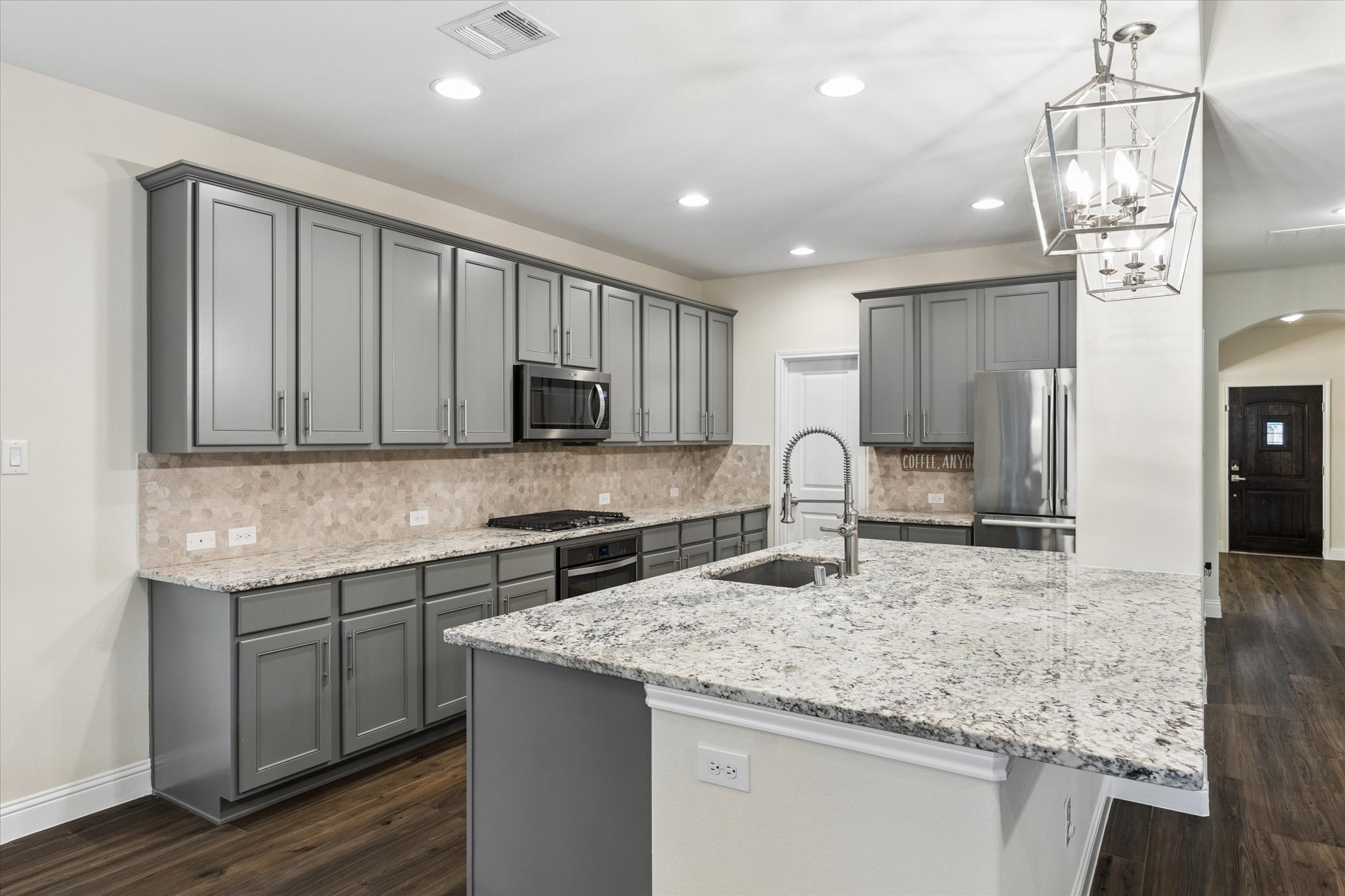 Beautifully updated kitchen featuring an expansive granite island with a farmhouse-style sink, abundant gray cabinetry, stainless steel appliances, and a decorative tile backsplash. Recessed lighting and stylish pendant fixtures create a bright, inviting atmosphere, while the generous counter space and open layout make this kitchen ideal for everyday living and entertaining.