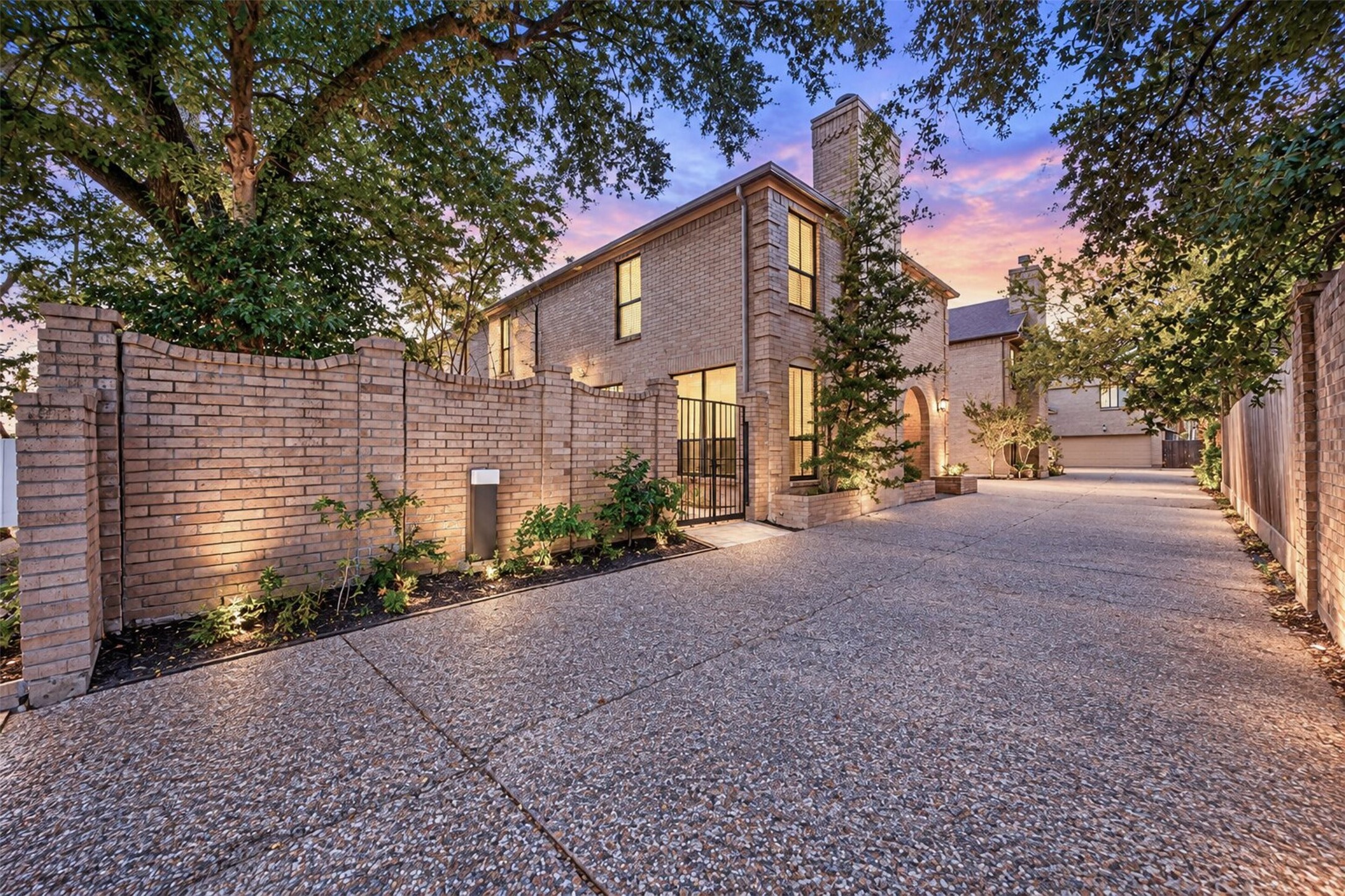 Classic brick exterior framed by mature trees, with a gated courtyard, arched entry and paved approach to the garage. Digitally enhanced twilight image.