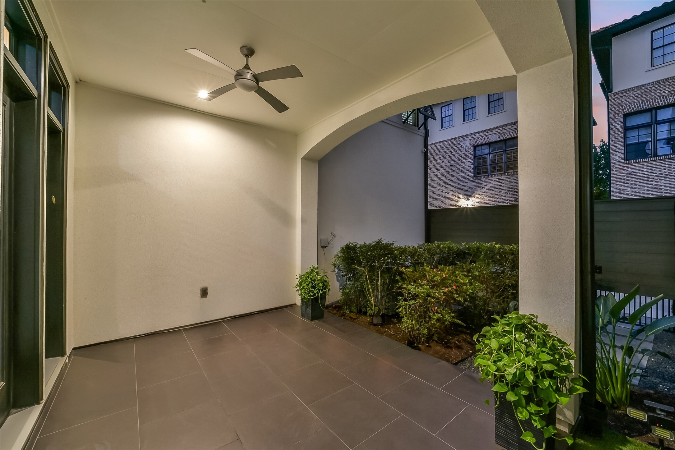 Private covered patio with an arched opening, ceiling fan, and large-format tile flooring, ideal for outdoor dining or relaxing year-round. Lush landscaping and a fenced courtyard setting add privacy, with French doors connecting seamlessly to the main living area.