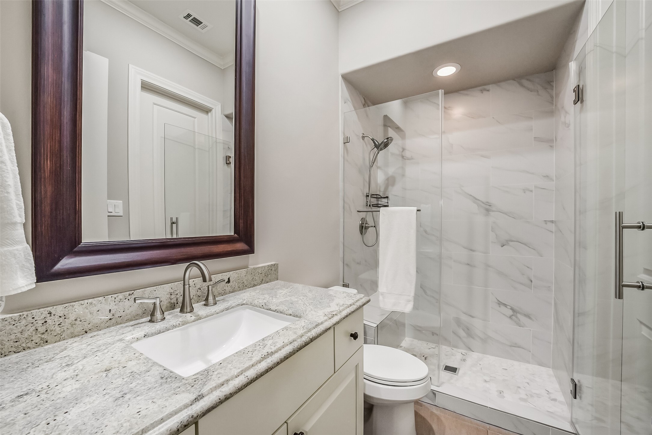 Secondary bath with a granite-top vanity, undermount sink, and a framed mirror. The walk-in shower has a frameless glass door, marble-look tile, a built-in bench, and a handheld shower head.