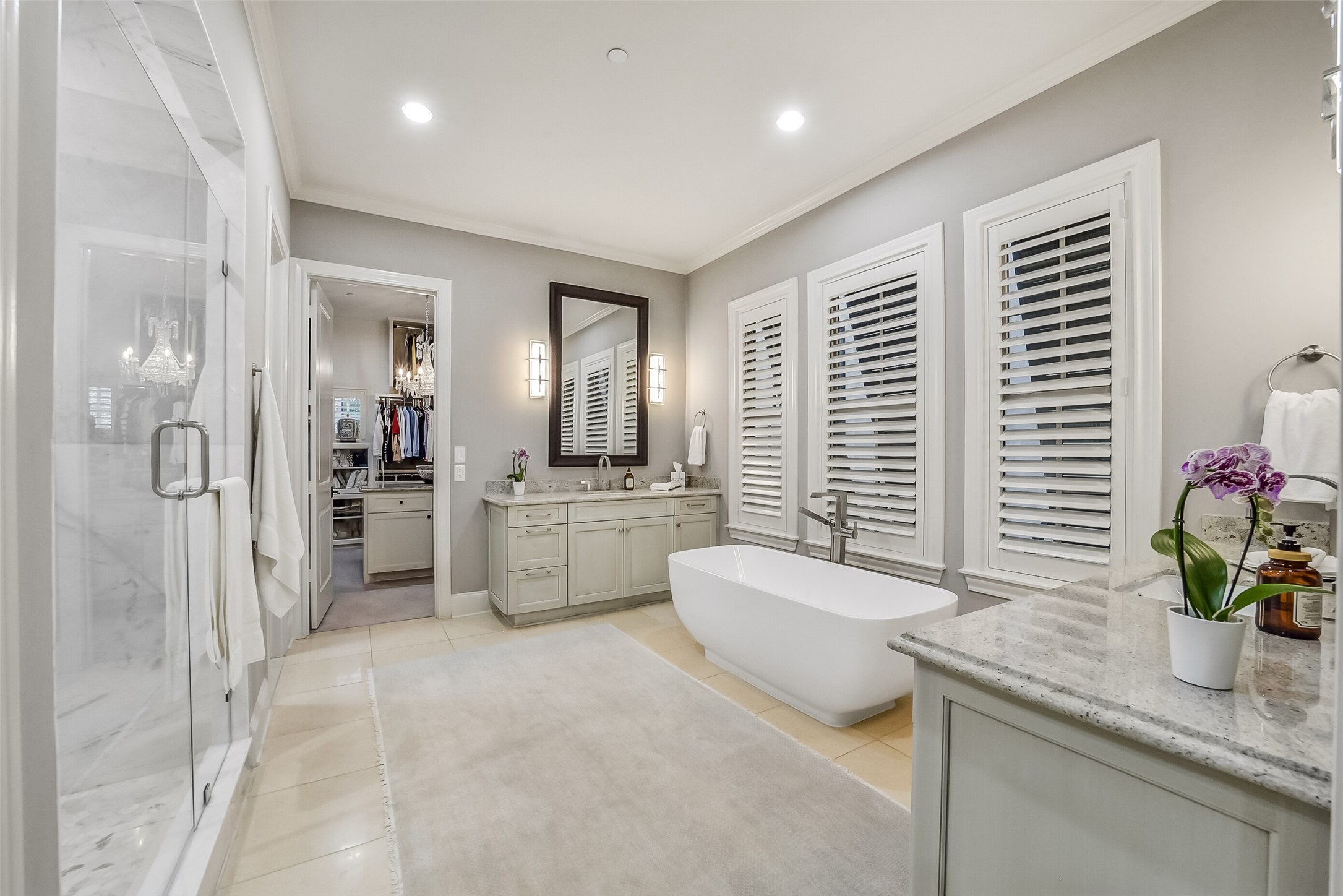 Luxurious primary bathroom featuring two separate vanities, a modern freestanding soaking tub framed by three shuttered windows for natural light and privacy, a seamless glass-enclosed shower, and direct access to 2 spacious walk-in closets.
