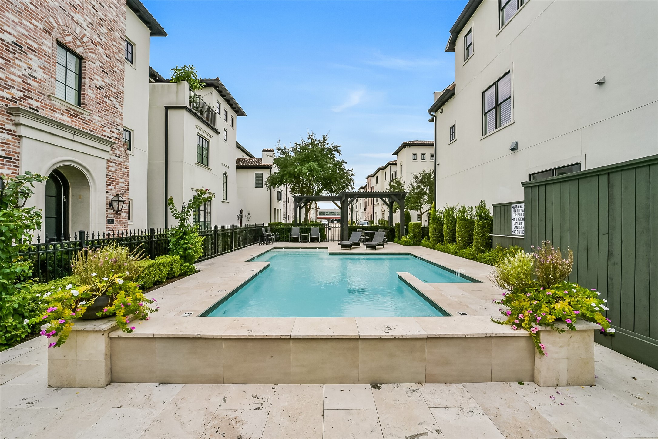 Resort-style community pool tucked inside the neighborhood, surrounded by travertine decking, lounge seating, and a shaded pergola. Wrought-iron fencing, lush landscaping, and colorful planters create a private, inviting retreat just steps from home.