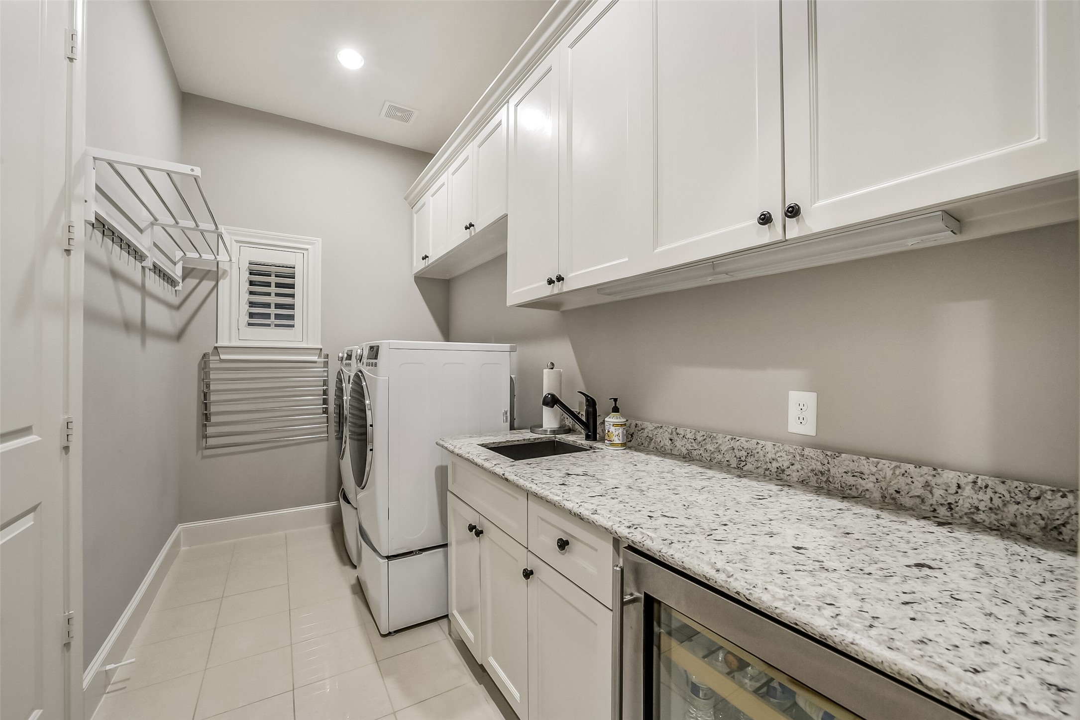 Well-appointed utility room with white cabinetry, granite countertops, and a built-in sink. Front-loading washer and dryer sit on pedestals, and wall-mounted drying racks and under-cabinet lighting make laundry day easier. A beverage cooler adds a nice extra.