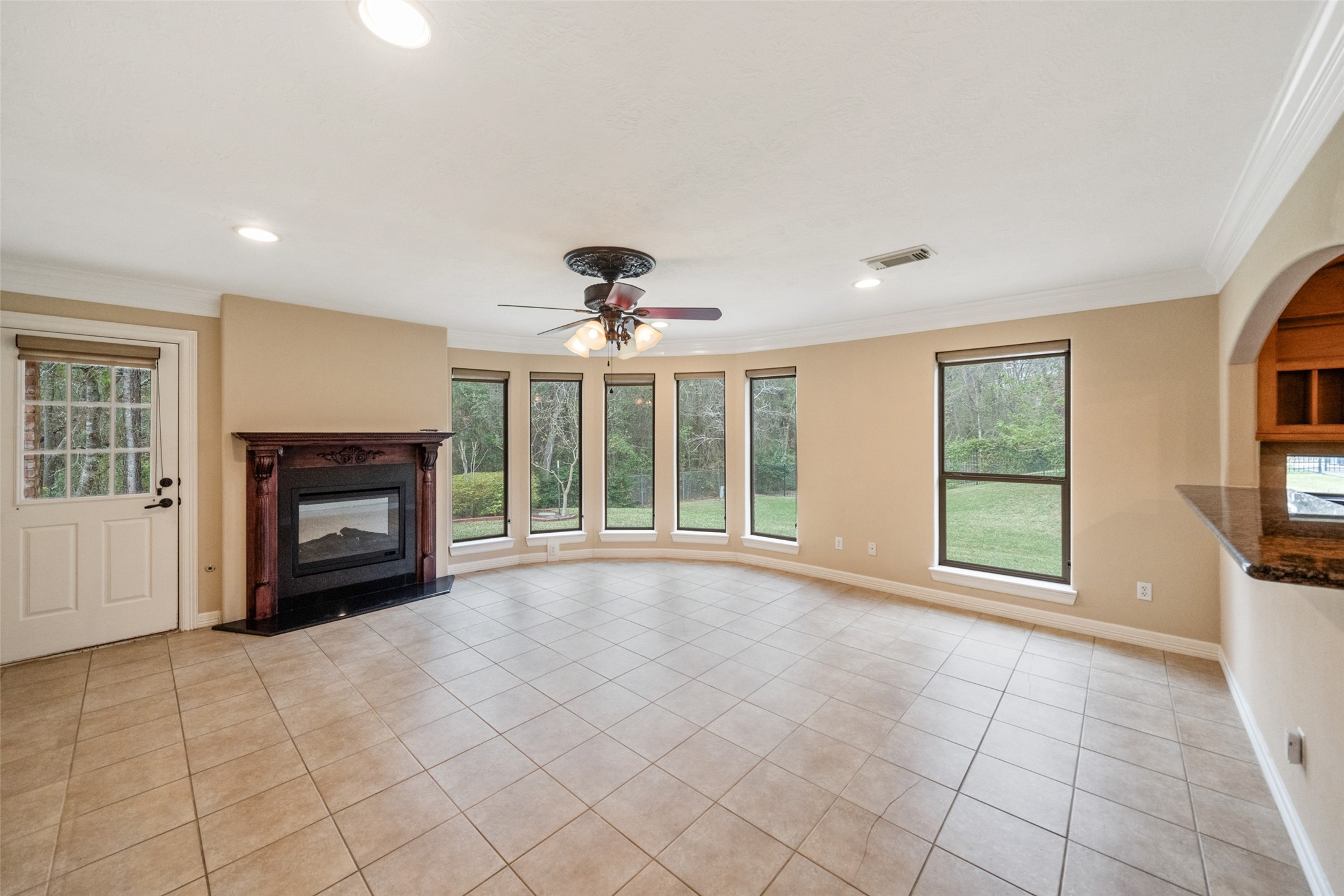 This spacious living room features a cozy fireplace, large windows offering plenty of natural light, tiled floors, and a ceiling fan. Its open design provides a welcoming and airy feel, perfect for relaxation or entertaining.