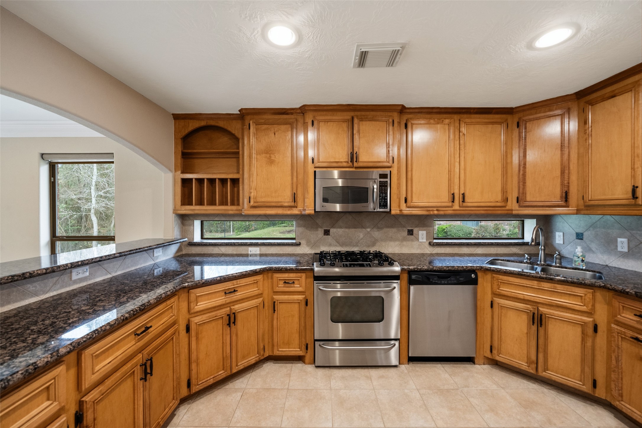 This kitchen features warm wood cabinetry, granite countertops, and stainless steel appliances, including a gas stove and dishwasher. It has a cozy corner sink with a window view, ample storage, and a tile backsplash, creating a functional and inviting space.