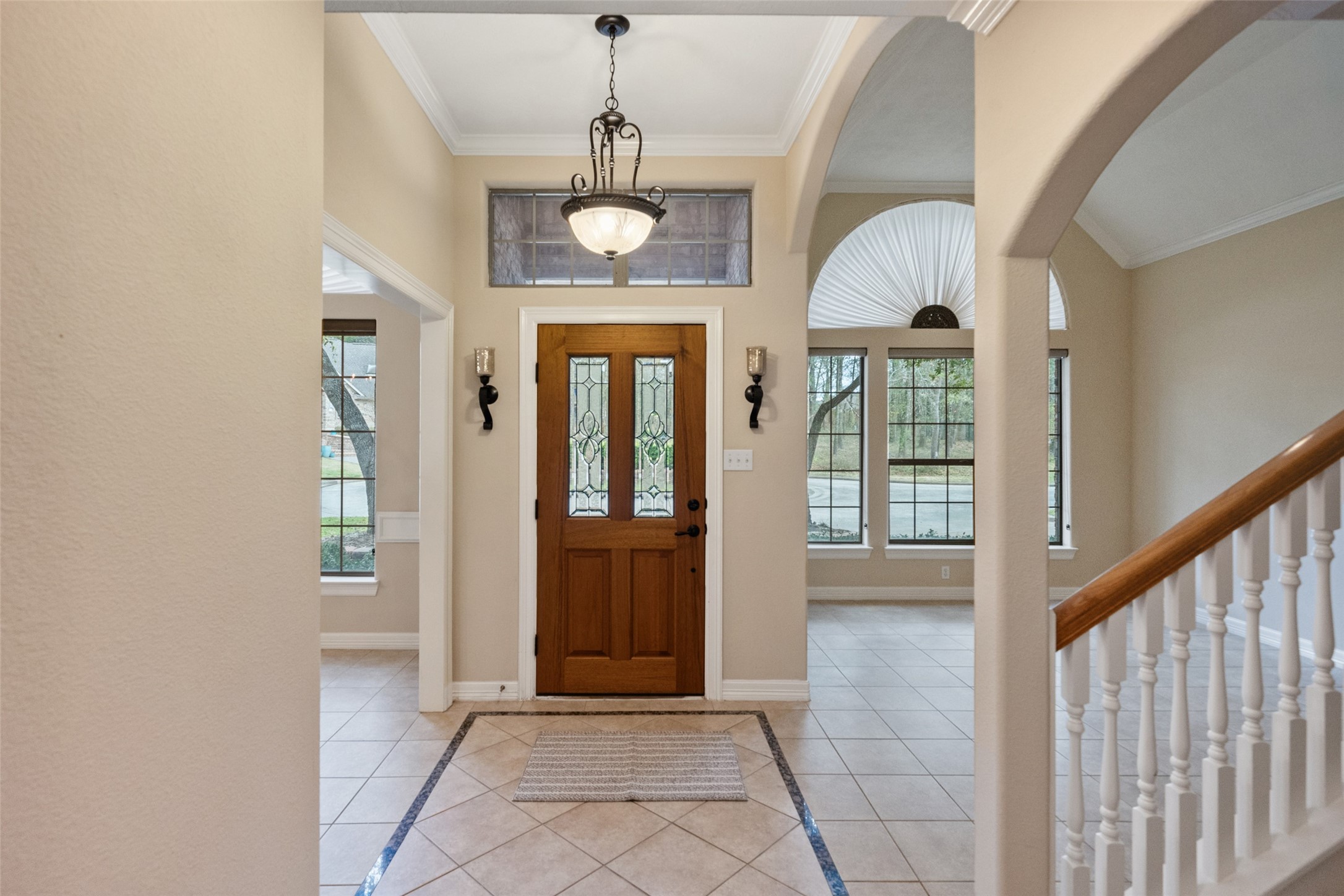 This inviting entryway features a wooden front door with decorative glass panels, elegant tile flooring, and a chandelier. Large windows bring in natural light, and the space opens up to a staircase with white railings, offering a welcoming and spacious feel. This inviting entryway features a wooden front door with decorative glass panels, elegant tile flooring, and a chandelier. Large windows bring in natural light, and the space opens up to a staircase with white railings, offering a welcoming and spacious feel.