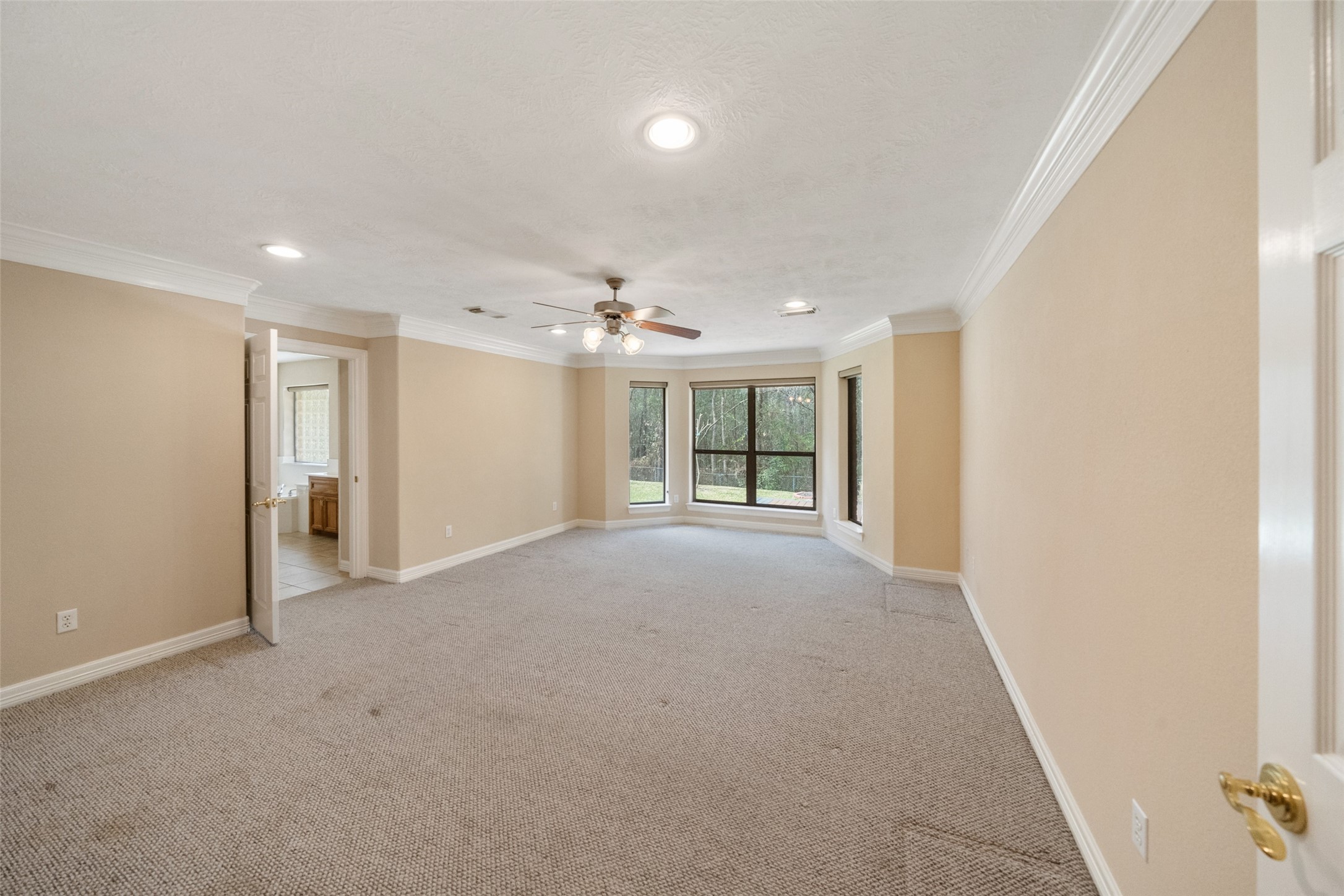 Spacious primary bedroom with crown molding. Features recessed lighting, ceiling fan, and large windows offering natural light. Adjacent to a bathroom with visible vanity area.