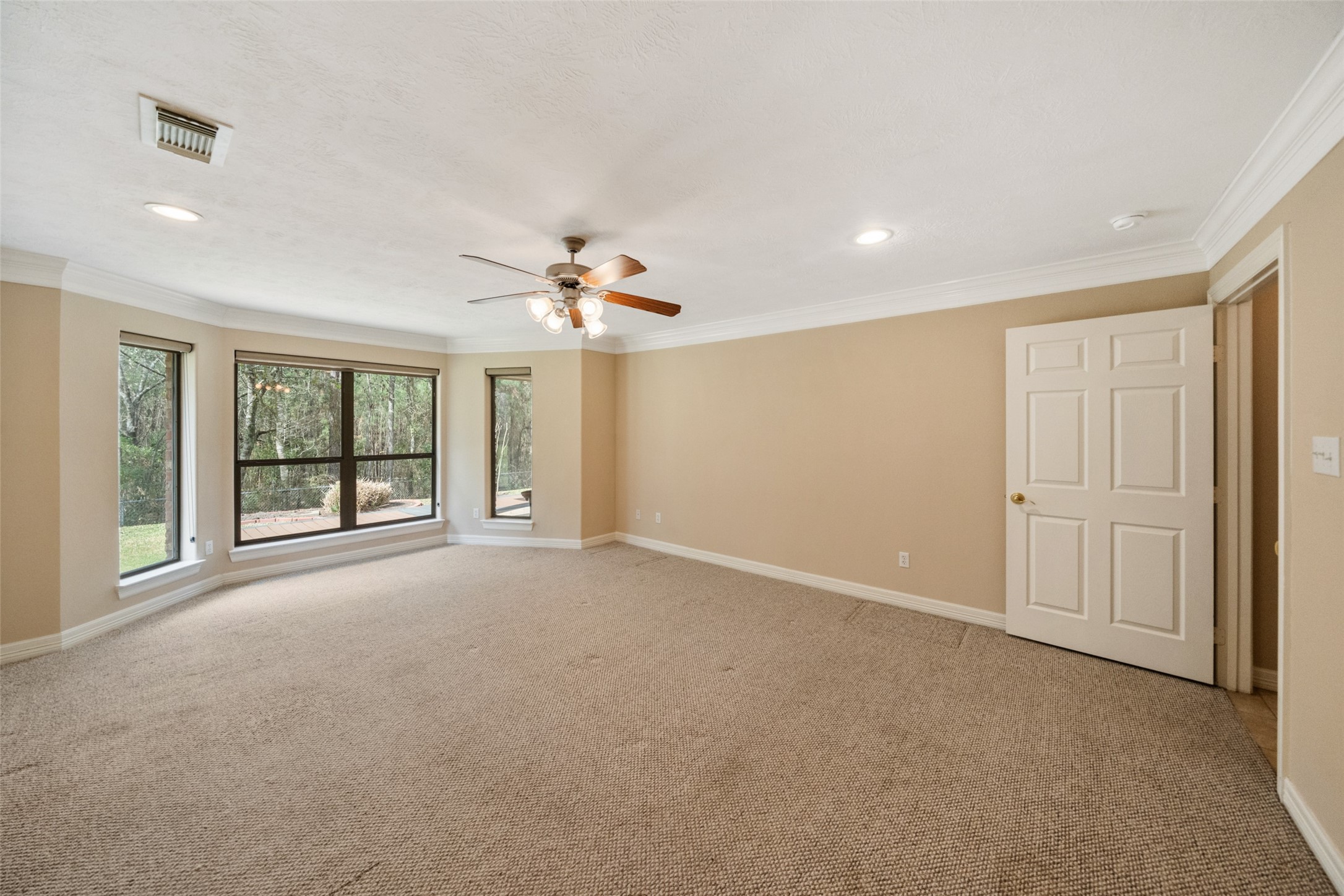 First floor primary bedroom features neutral walls and carpet, large windows with views of greenery, and a ceiling fan with recess lights, creating a bright and inviting atmosphere.