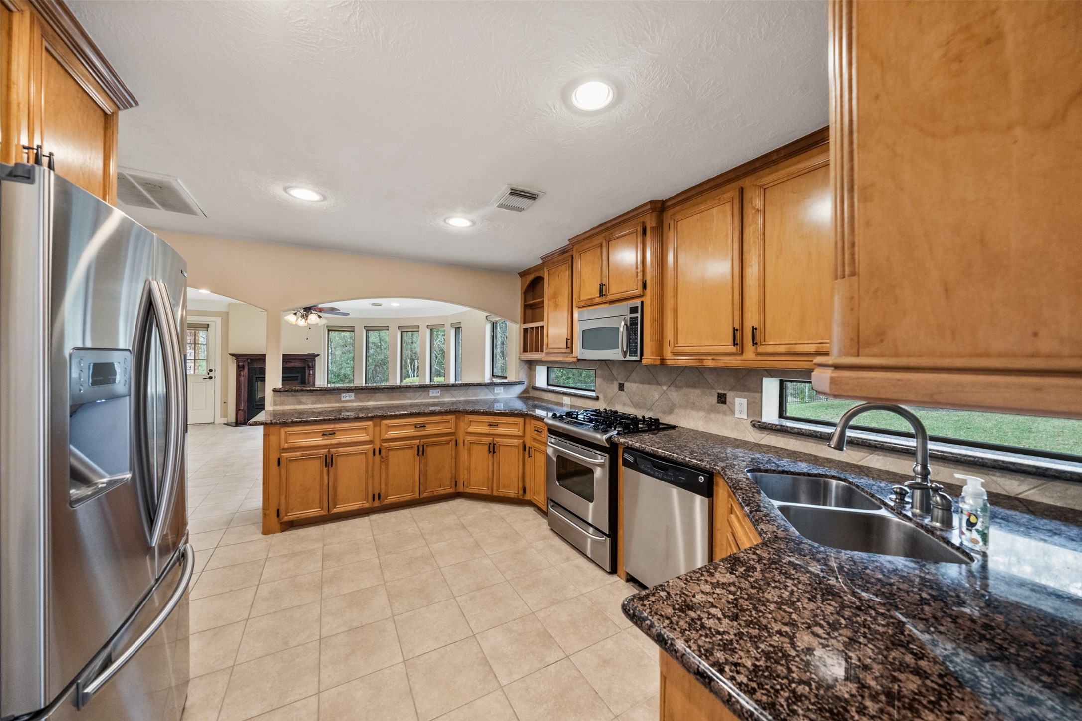 Spacious kitchen with granite countertops, stainless steel appliances, and ample wooden cabinetry. It features a double sink, gas stove, and a view into a bright living area with large windows.