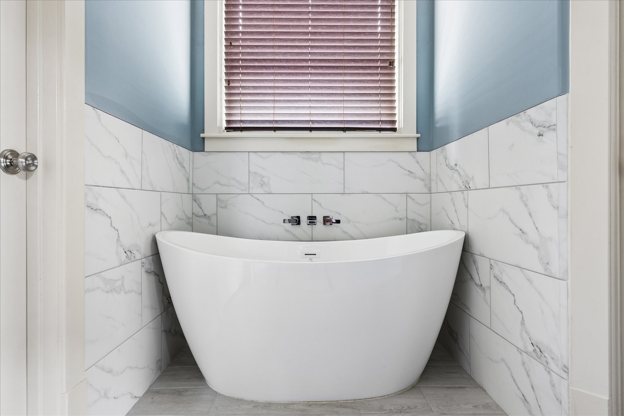 A sculptural freestanding soaking tub takes center stage beneath the window, framed by soft blue walls and elegant tile.