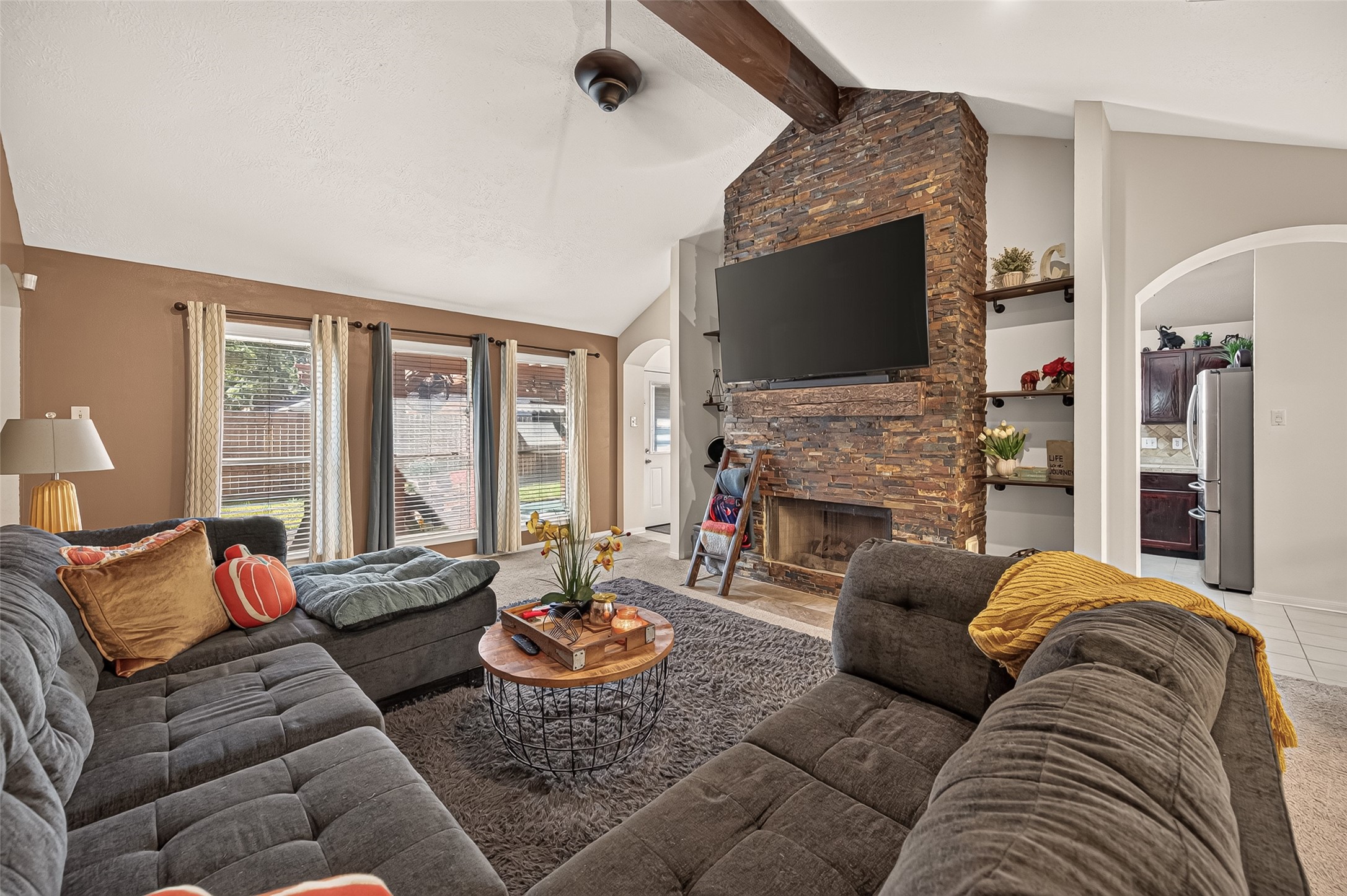 Spacious living room featuring a vaulted ceiling with a rustic beam, a striking stone fireplace, and large windows that fill the space with natural light.