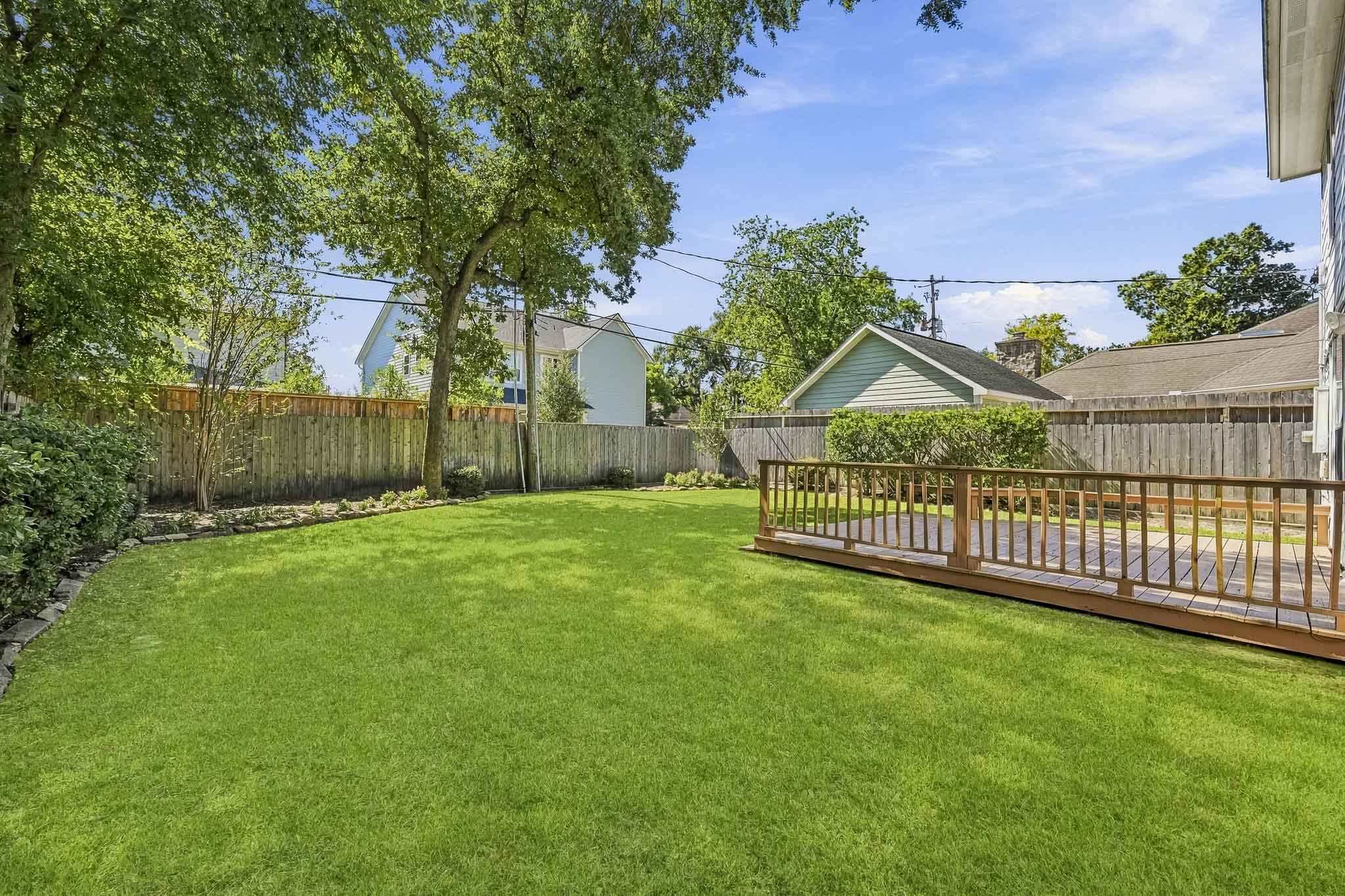 Expansive, fully fenced backyard with mature shade trees, lush lawn, and a large wooden deck for outdoor entertaining.