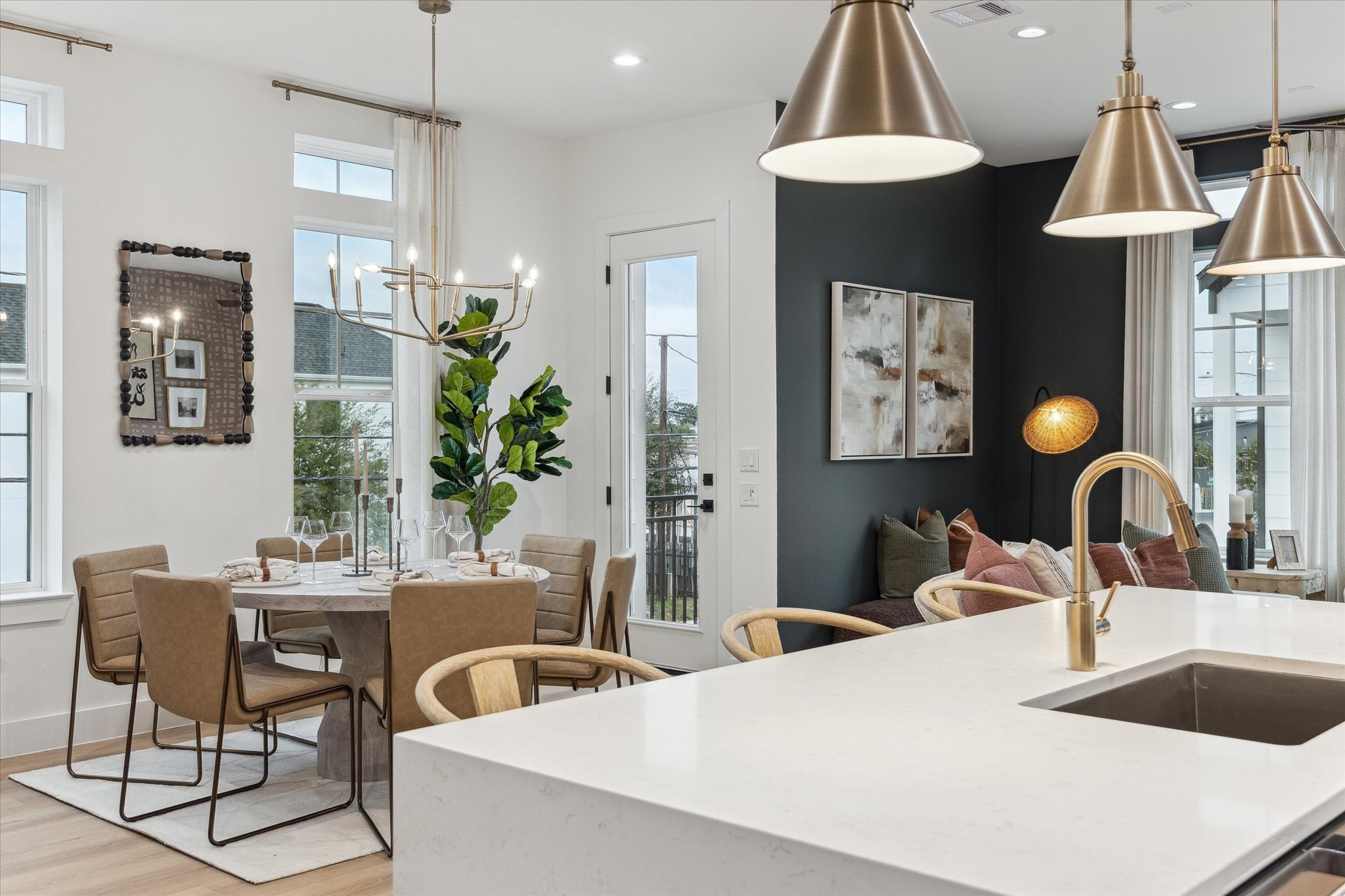 Elegant open-concept dining and living area featuring a modern kitchen island with gold fixtures, chic pendant lighting, and a cozy seating nook. Large windows and a stylish chandelier enhance the airy ambiance.