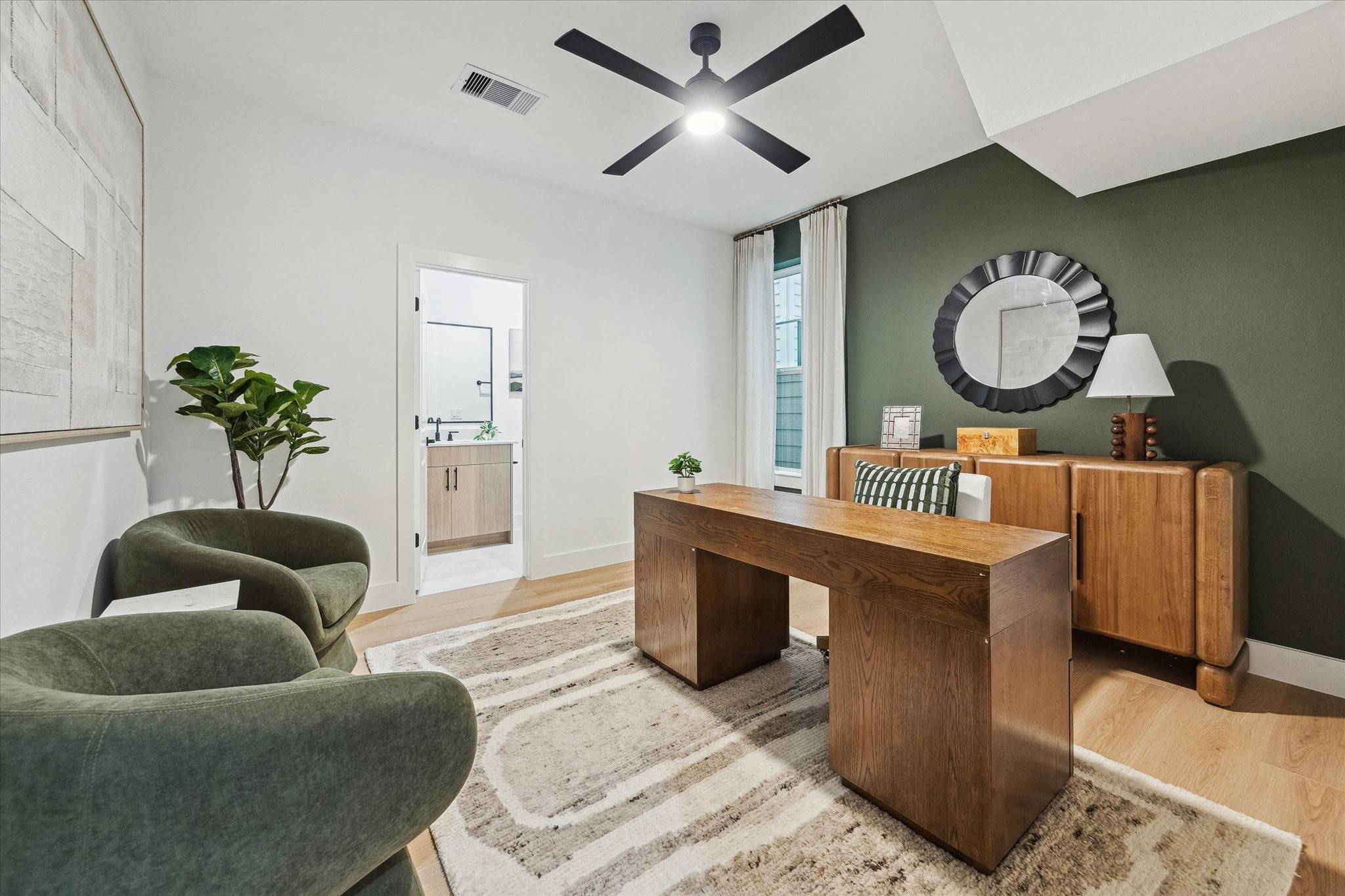 Chic home office with a modern wood desk, cozy green armchairs, and stylish decor. A dark accent wall complements the room's contemporary design, complete with a ceiling fan and ample natural light.