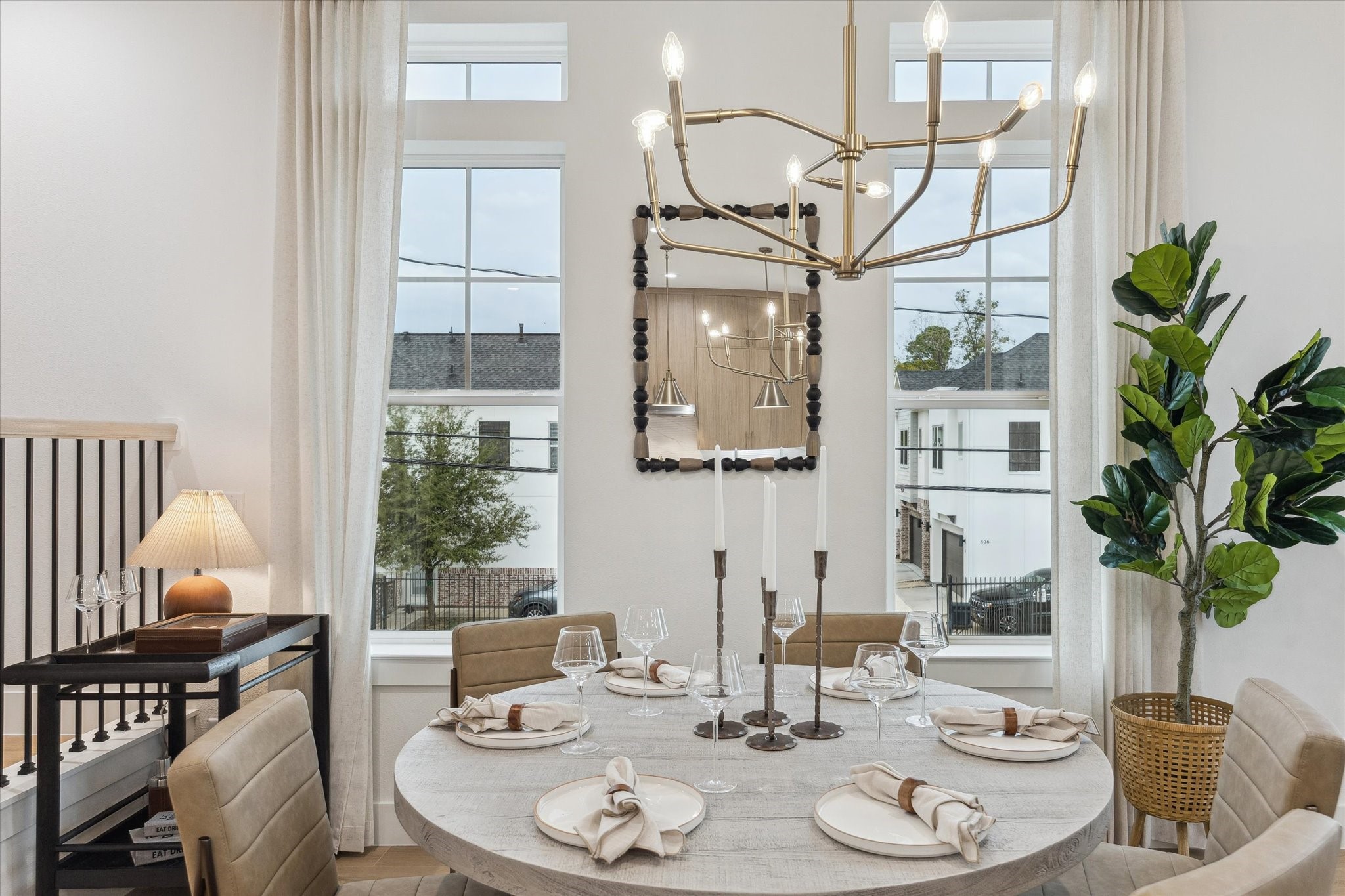 Elegant dining area featuring a round table set for four, under a striking modern chandelier. Large windows provide ample natural light, complemented by tasteful decor and a lush potted plant for a welcoming ambiance.