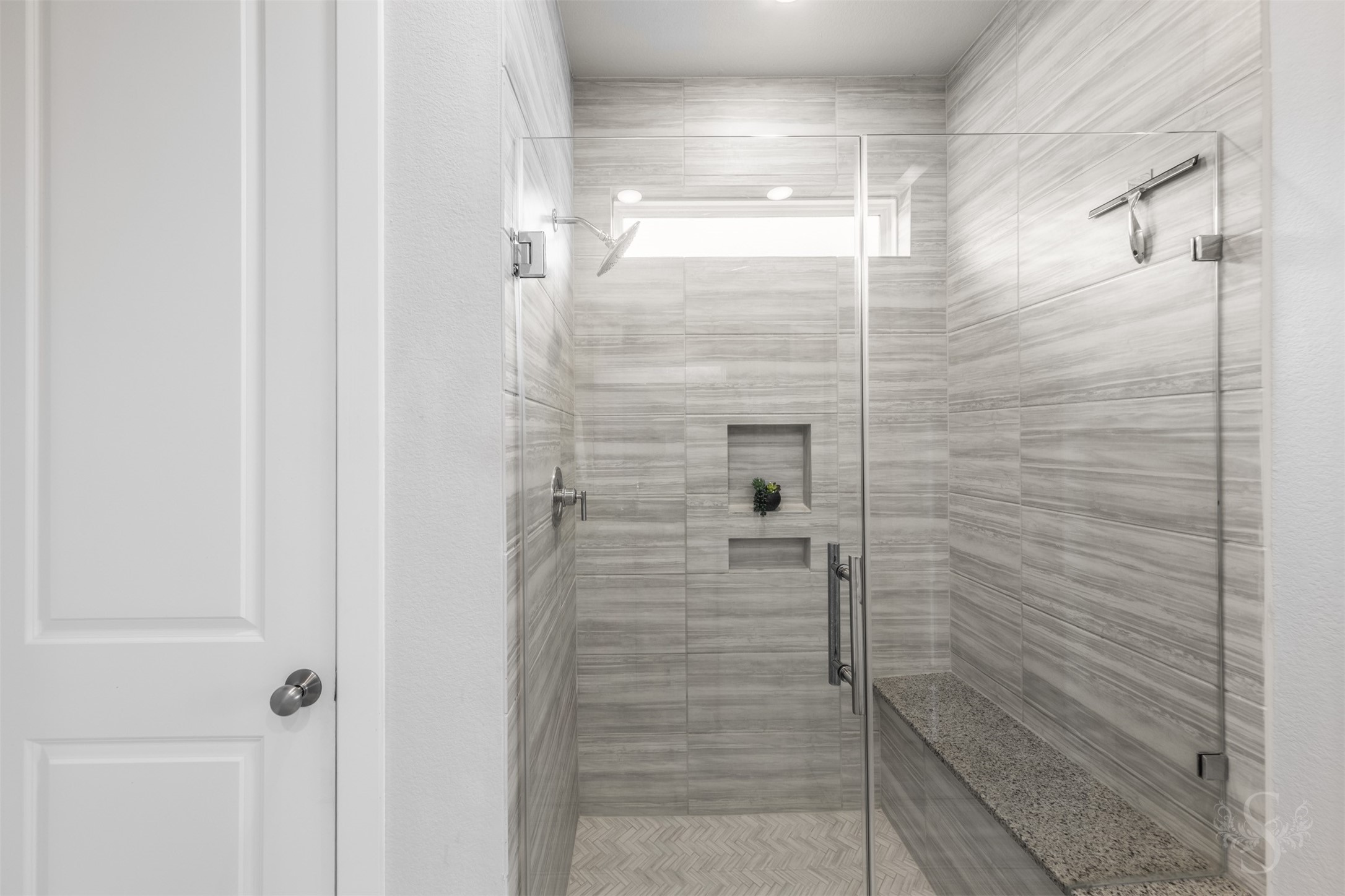 The luxurious walk-in shower showcases a frameless glass enclosure, beautiful tilework and a built-in bench. Recessed niches provide convenient storage, while a high window welcomes daylight into this relaxing space.