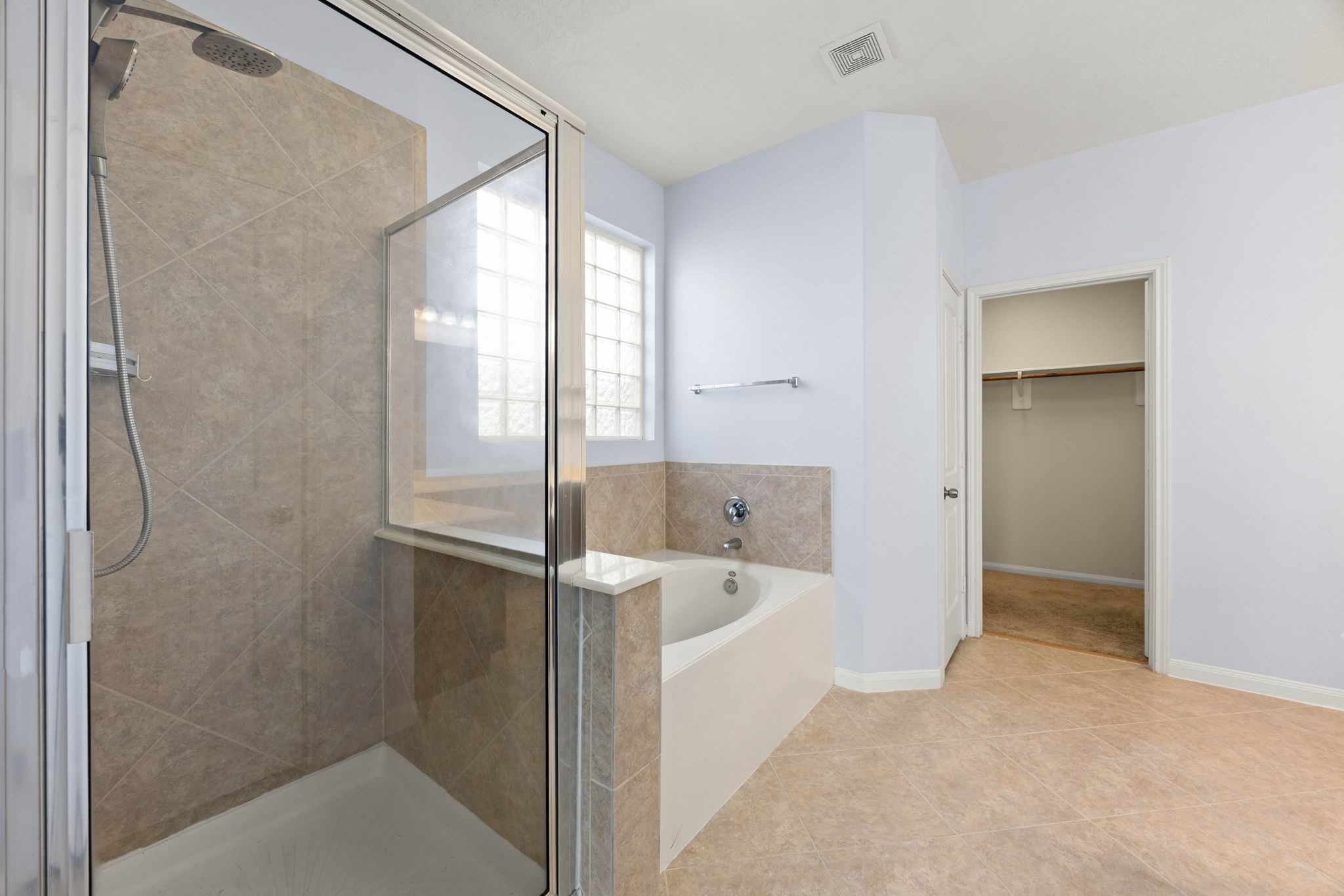 A luxurious spa-like retreat awaits in the primary en-suite, featuring a separate deep soaking tub and a dedicated glass-enclosed walk-in shower. The spaces are elegantly tied together with neutral diagonal tile surrounds, a privacy-enhancing glass block window that lets in soft natural light, and direct access to a spacious walk-in closet just steps away.