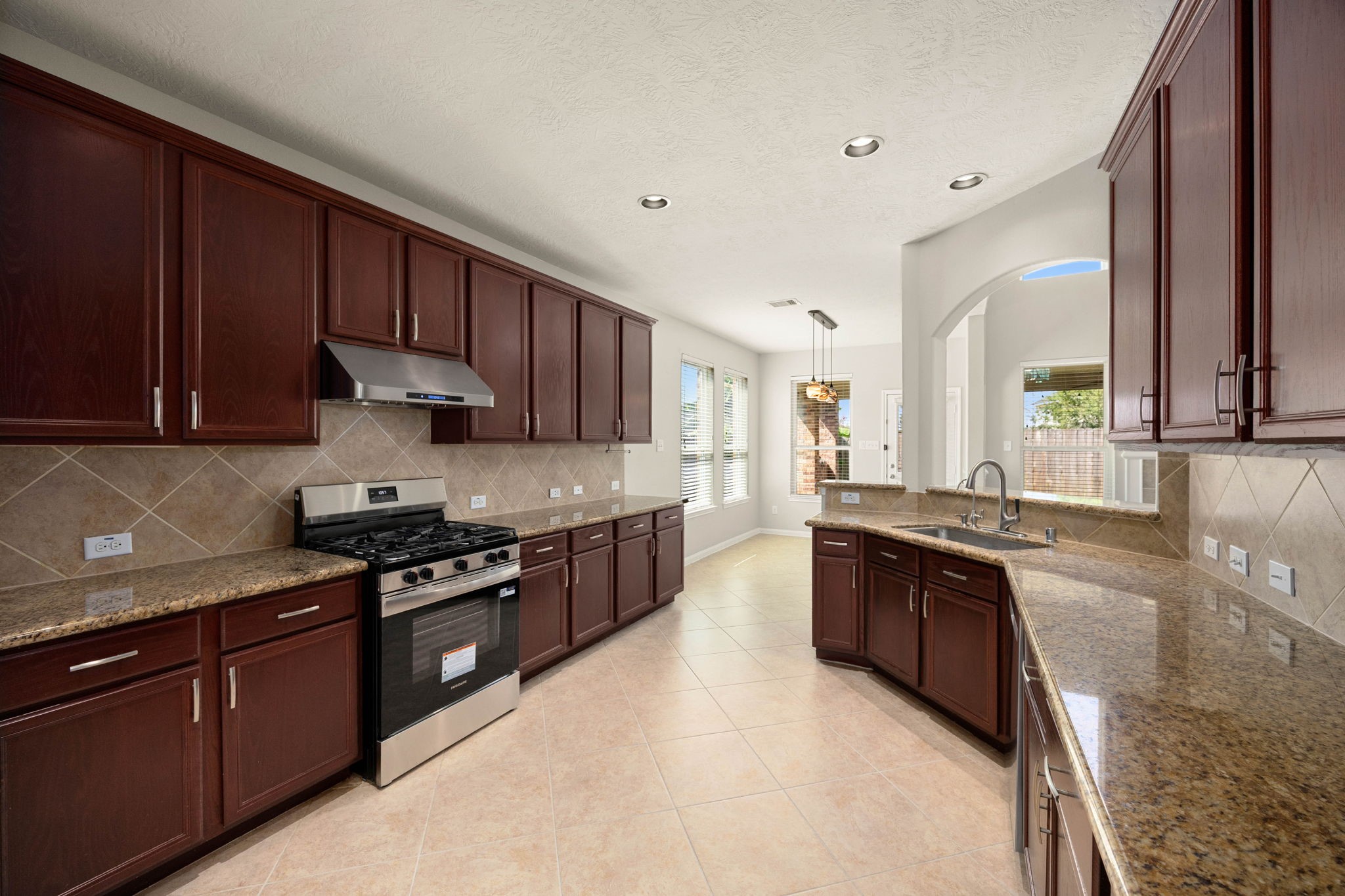 This reverse angle of the spacious kitchen highlights the impressive length of the granite countertops and the extensive cabinetry layout. The view emphasizes the integrated flow from the cooking zonefeaturing a stainless steel gas range and matching vent hoodstraight into the sunlit breakfast nook and out toward the backyard, making it an ideal setup for hosting and daily meal prep.