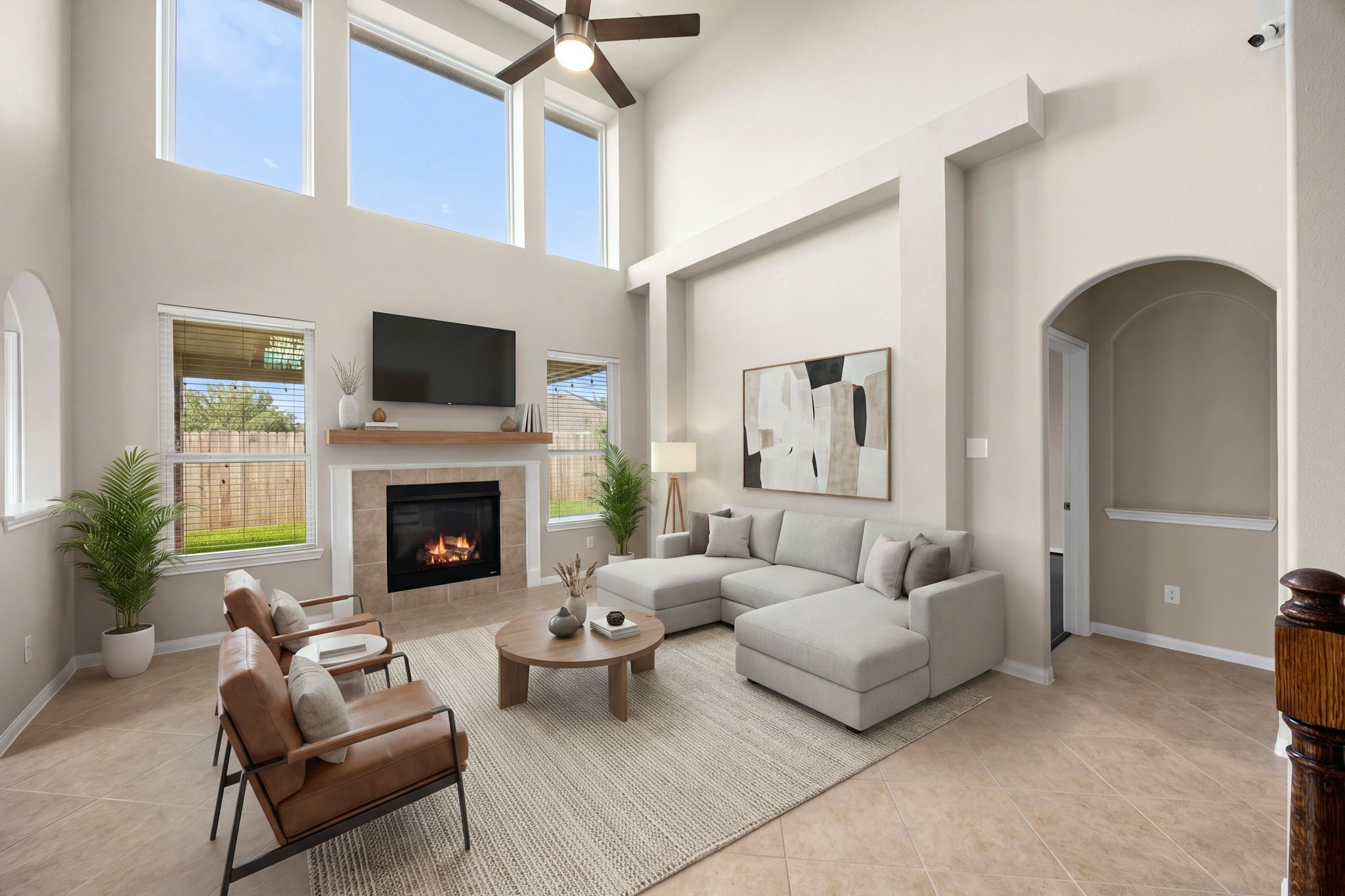 Soaring two-story ceilings and an impressive wall of upper windows define this spectacular living room, filling the space with an exceptional amount of natural light. A cozy fireplace with a custom wood mantel serves as the room's focal point, beautifully complemented by neutral tile flooring, a modern ceiling fan, and elegant architectural arches that transition seamlessly into adjacent living spaces. *Virtually Staged
